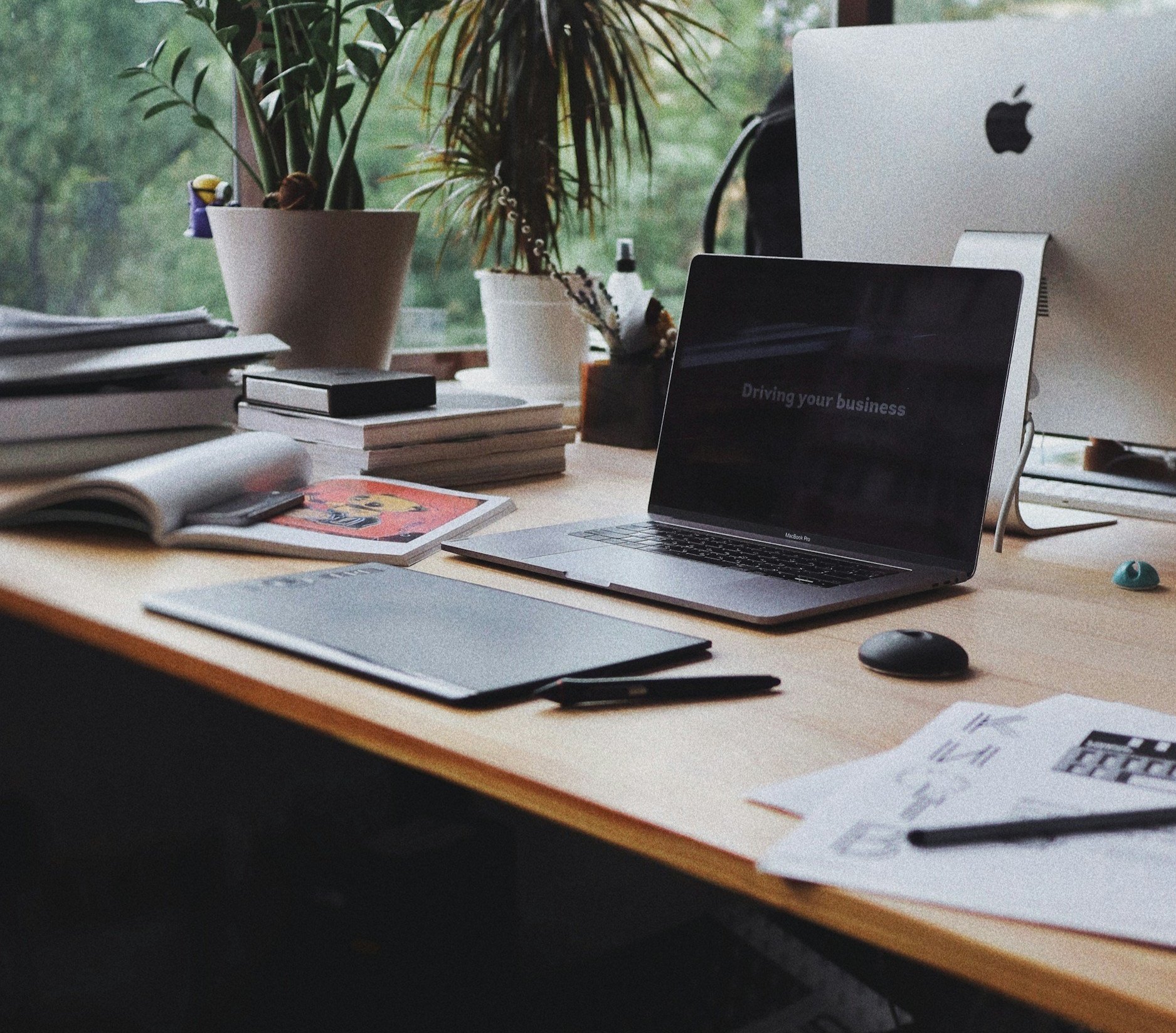 デスクの上にノートパソコン、マウス、ペン、書類、本、ペンタブレット、2つの植物の鉢置きが置かれています。背景には窓と外の緑の景色が見えます。