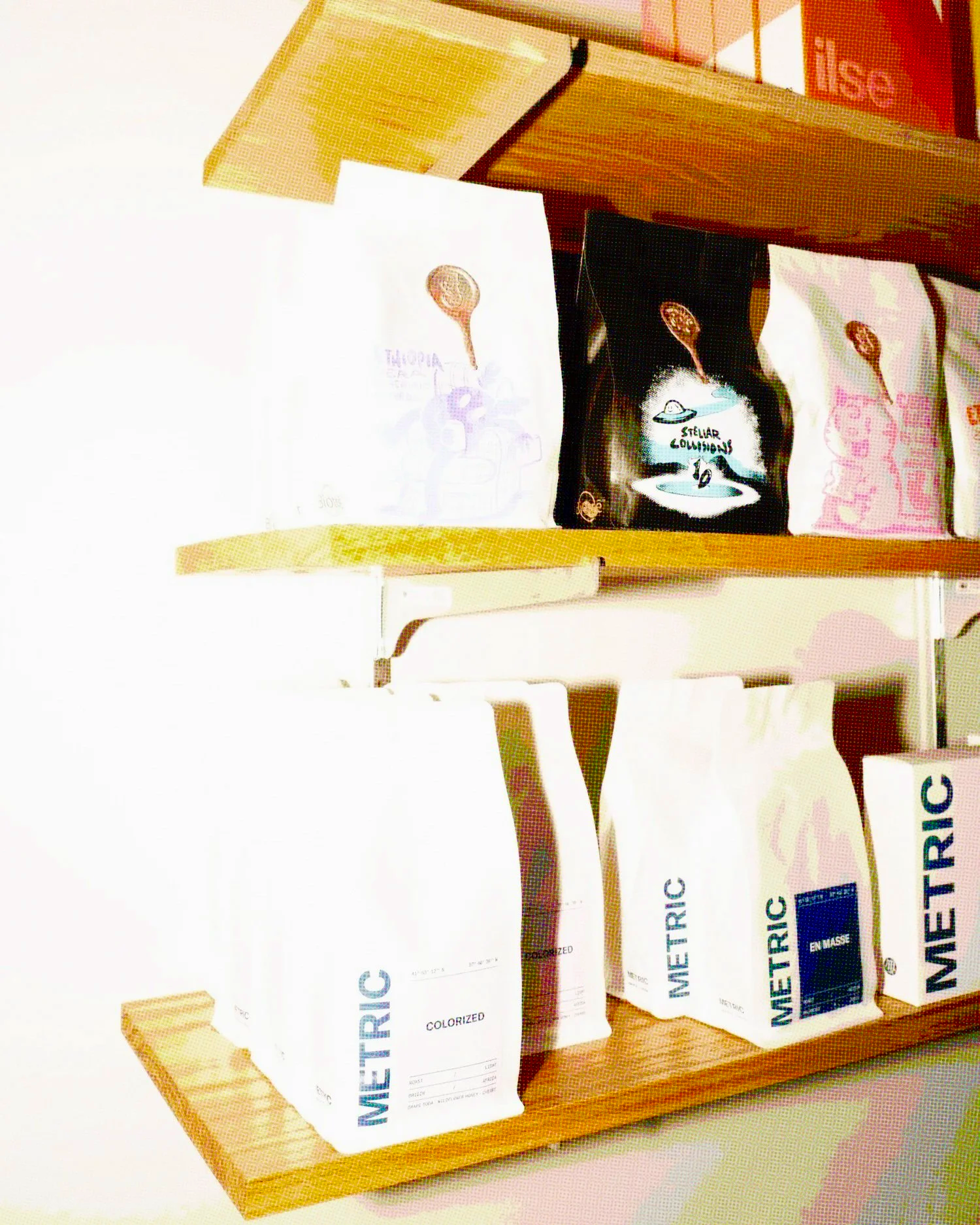 Wooden shelf displaying bags of METRIC coffee and other specialty coffee roasters. A black bag with UFO and 'Stellar Collisions' text