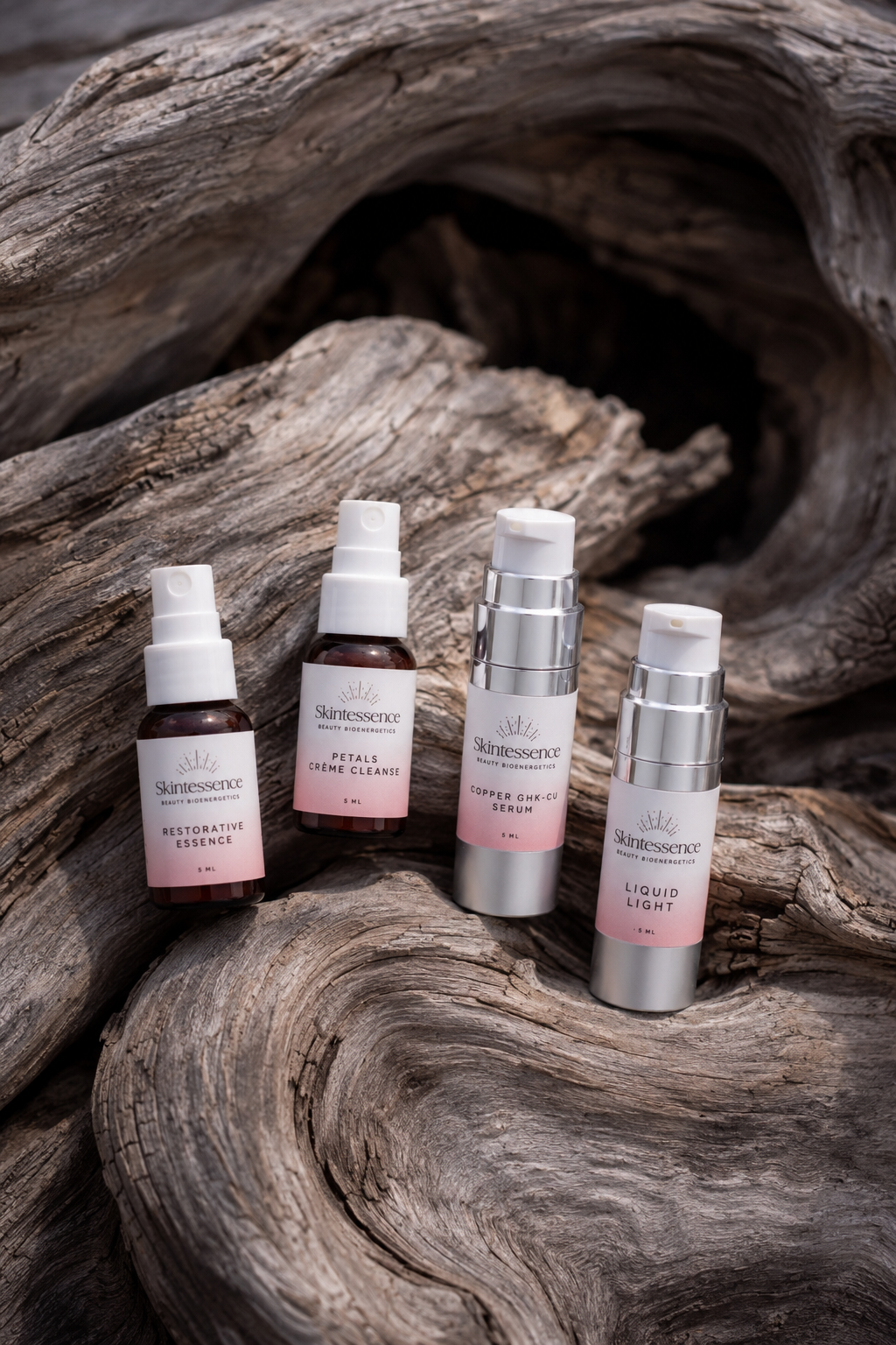 Four skincare bottles placed on weathered driftwood, featuring products from Skintessence with pink and silver labels.