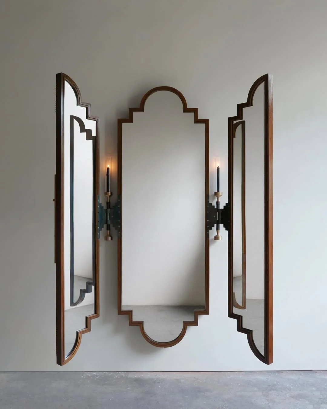 Three ornate wooden mirrors with decorative frames mounted on a white wall, with two wall-mounted candle sconces between the outer mirrors.