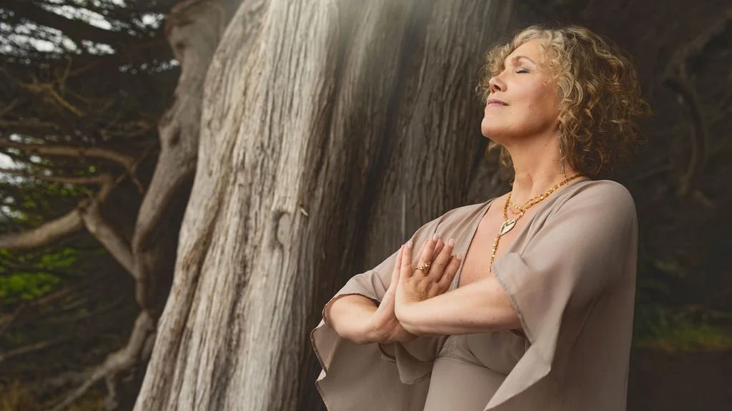 A woman with curly blonde hair practicing yoga outdoors next to a large tree with closed eyes and hands in a prayer position.