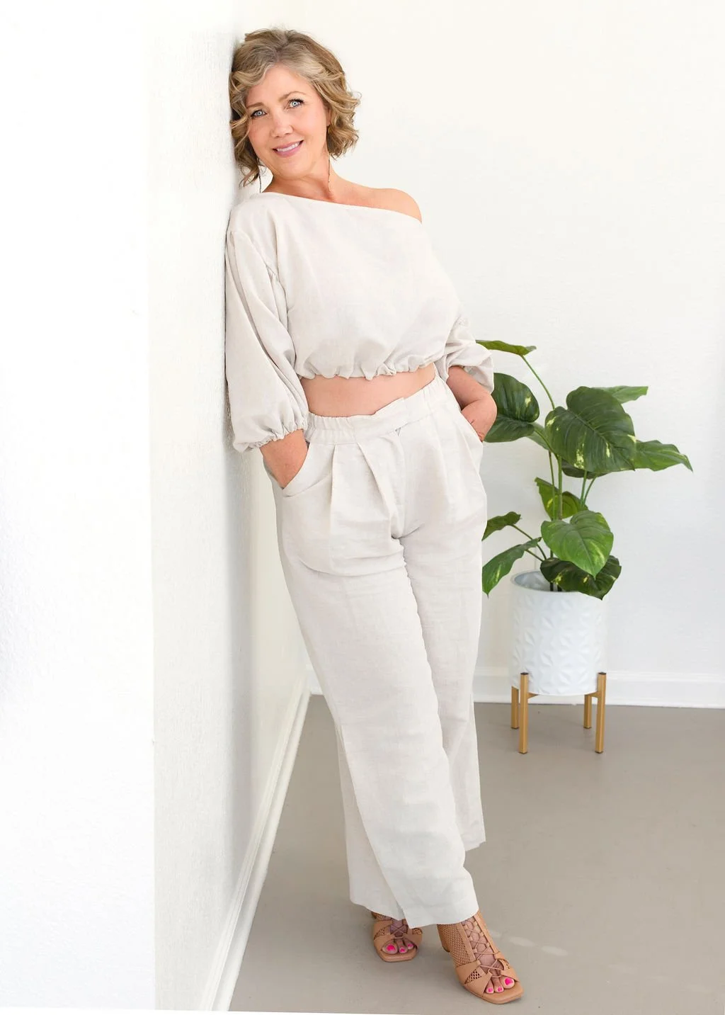 A woman with blonde, curly hair in a beige outfit, leaning against a white wall, with a green potted plant nearby.
