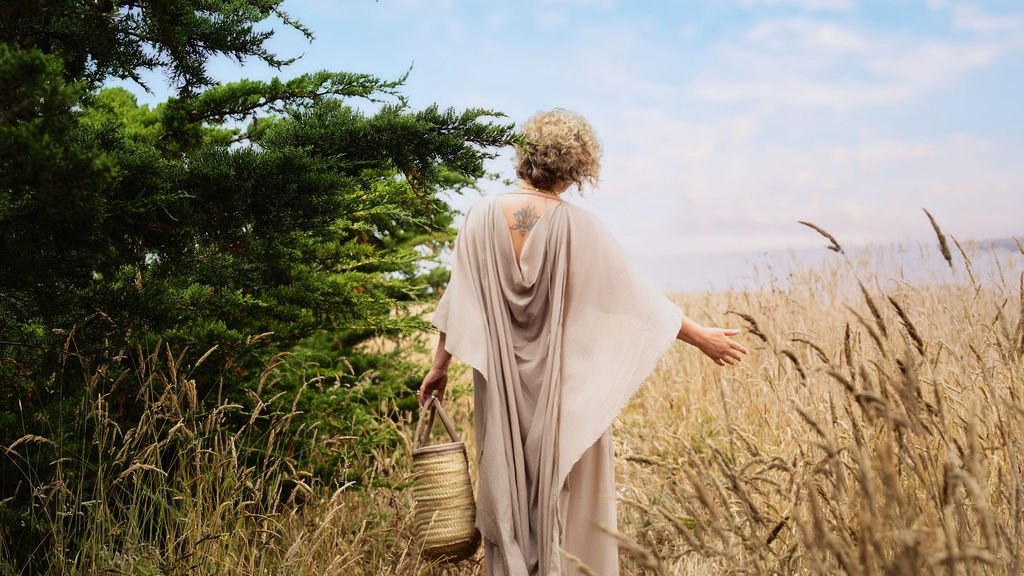 A woman with curly blonde hair, wearing a flowing beige dress, carrying a basket, walking through a grassy field with a tree nearby, on a partly cloudy day.