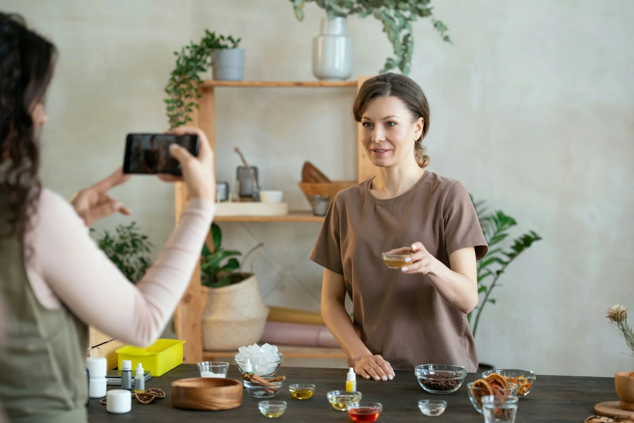 Woman with short hair and a brown shirt holding a glass of tea, being photographed by another person in a setting with plants and various items on a table.