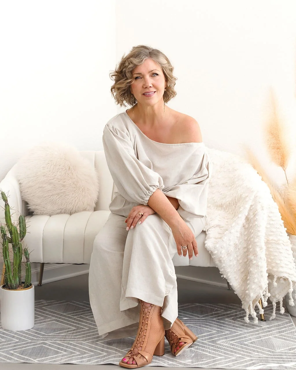 A woman with curly blonde hair wearing a beige off-the-shoulder top and matching pants, sitting on a white sofa in a neutral-toned room.