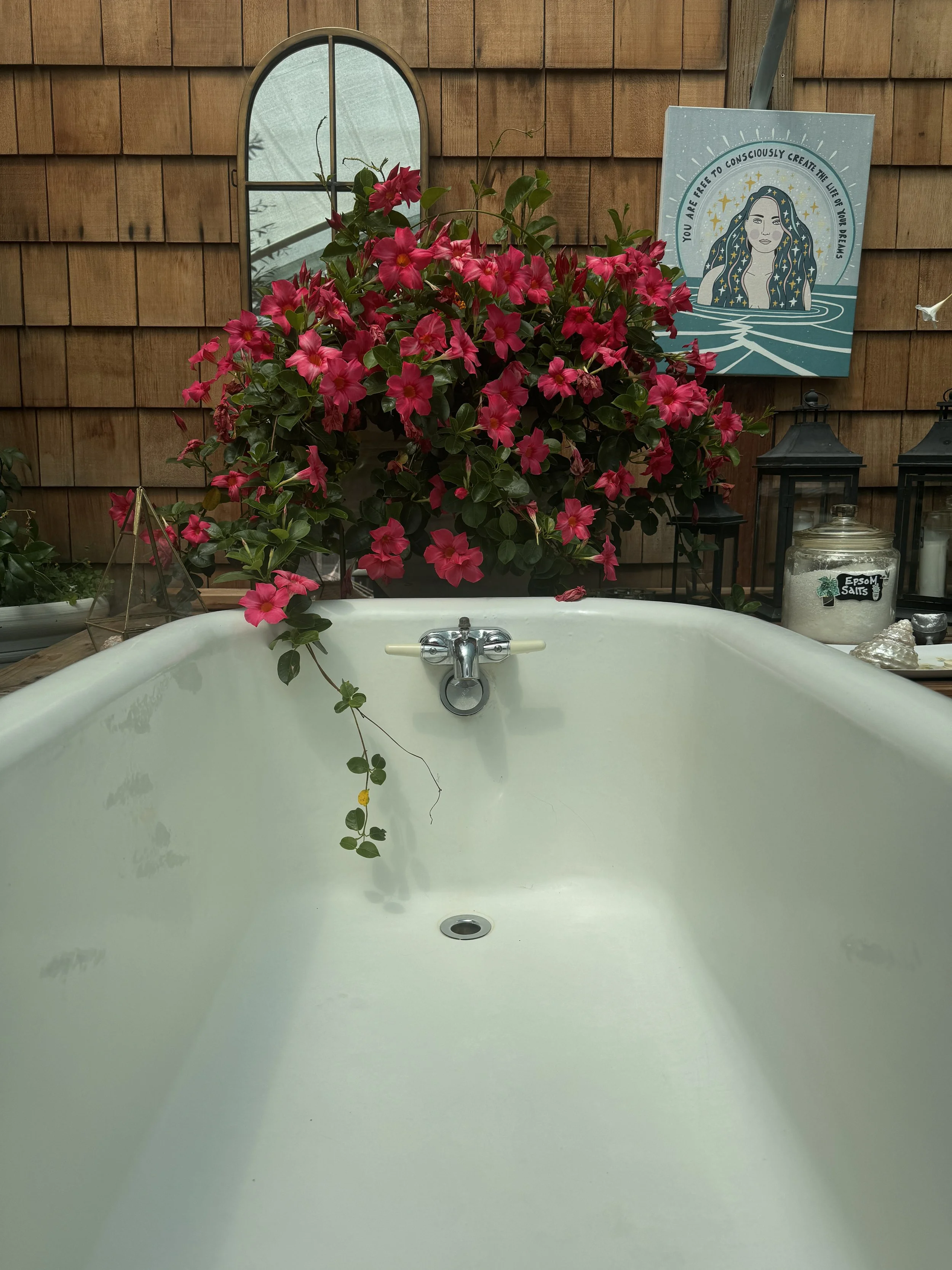 A white bathtub with a chrome faucet showing a cluster of pink flowers and green leaves overflowing into it. Behind, there is a wooden wall with a small arched window, a blueprint-style illustration with a woman and celestial theme, two black lanterns, and a jar of Epsom salts.