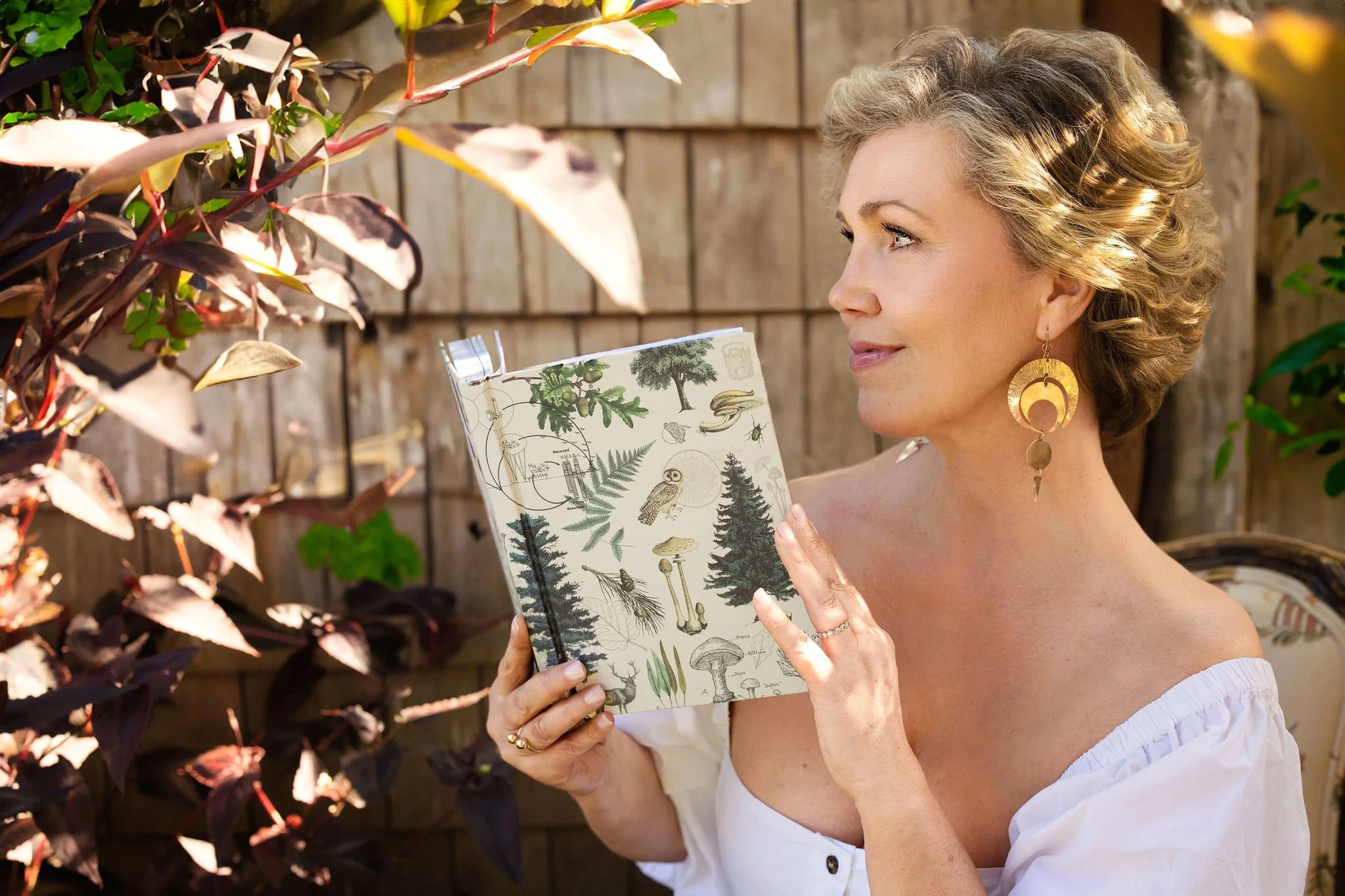 A woman with curly blonde hair and large gold earrings is outside, holding an illustrated book about plants and fungi while standing in front of a wooden fence and leafy plants.