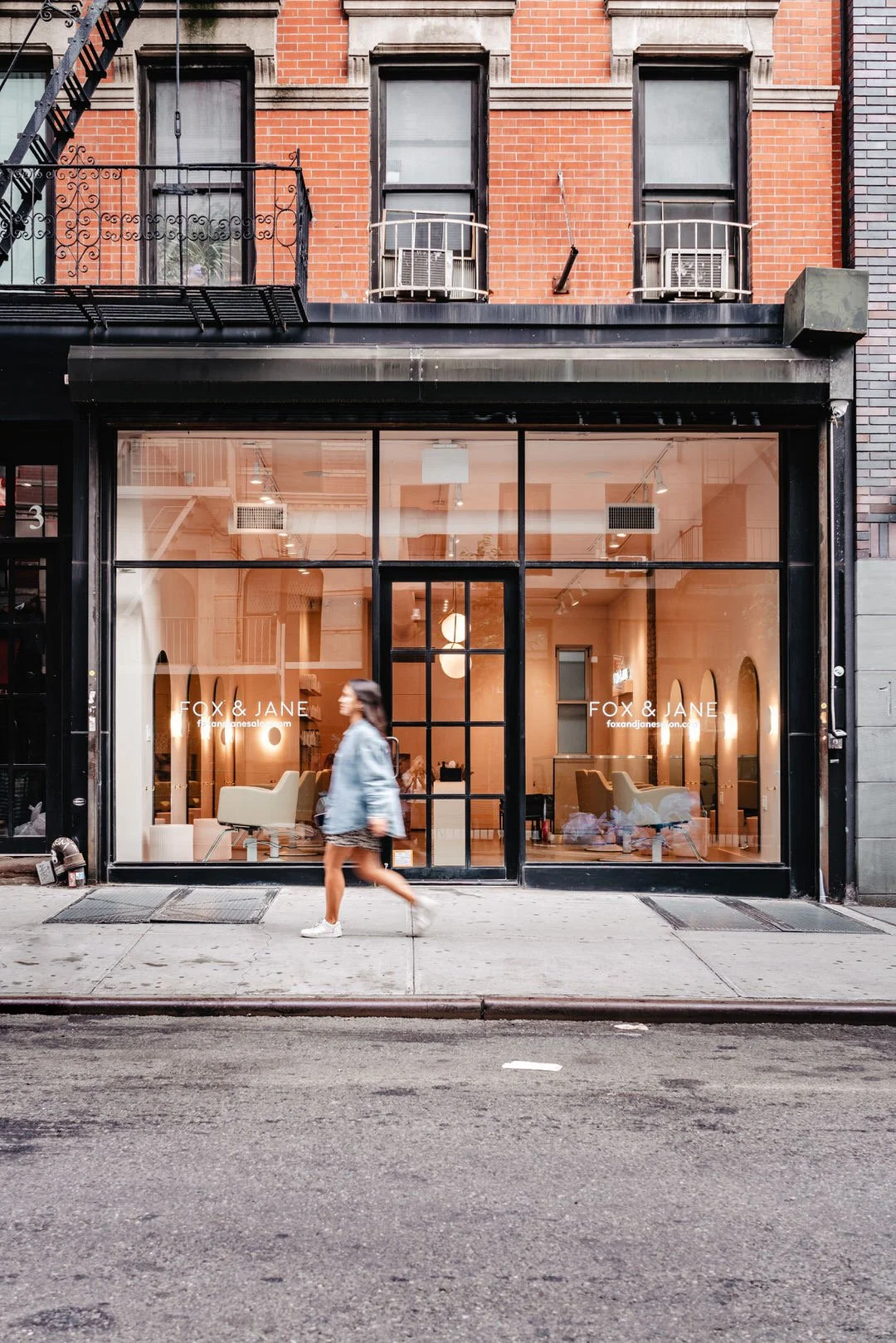 The storefront is a modern hair salon named 'FOX & JANE' with large glass windows showing a cozy interior with white chairs and warm lighting. A woman in a denim jacket and shorts is walking past on the sidewalk in front of the salon, on a city street with brick buildings.