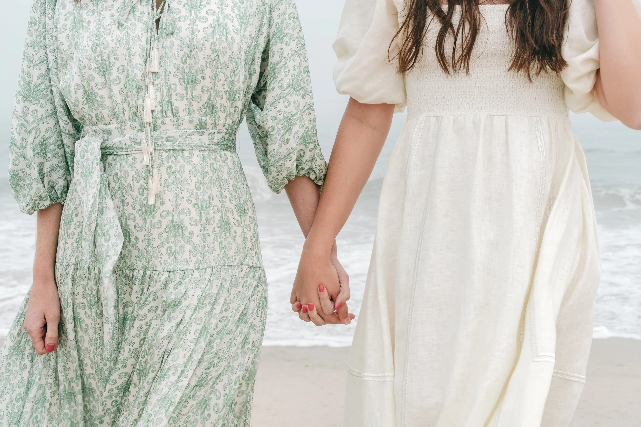Two women holding hands on the beach, wearing light-colored dresses for a apparel company brand shoot in San Diego.
