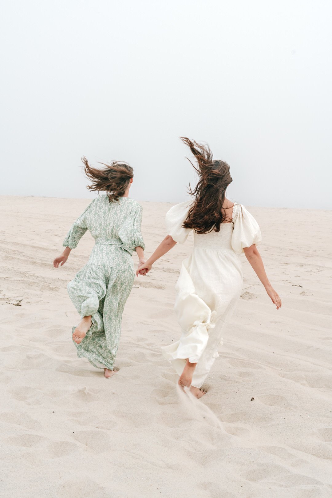 Two women wearing white and light-colored dresses running hand-in-hand on a sandy beach with wind-blown hair.