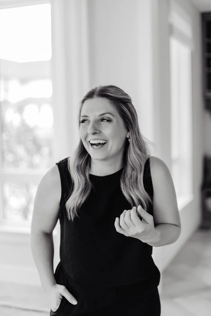 A woman with long wavy hair, wearing a sleeveless black top, smiling and laughing while standing indoors near a window.
