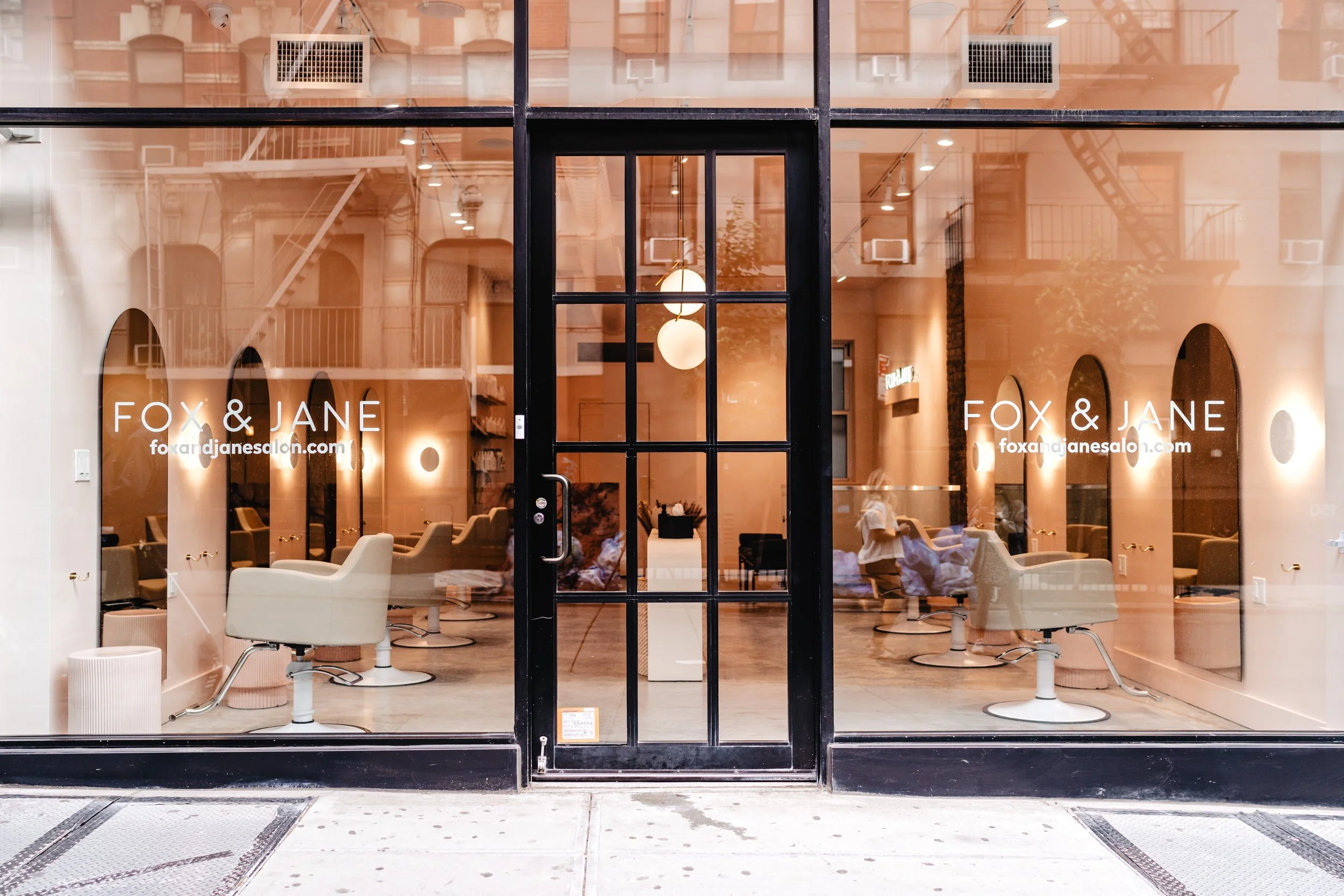 Front view of a modern salon with glass storefront, black frame door, and white signage reading "FOX & JANE" for their salon branding photoshoot. Inside, cream-colored salon chairs, round ceiling lights, and a staffed environment are visible.