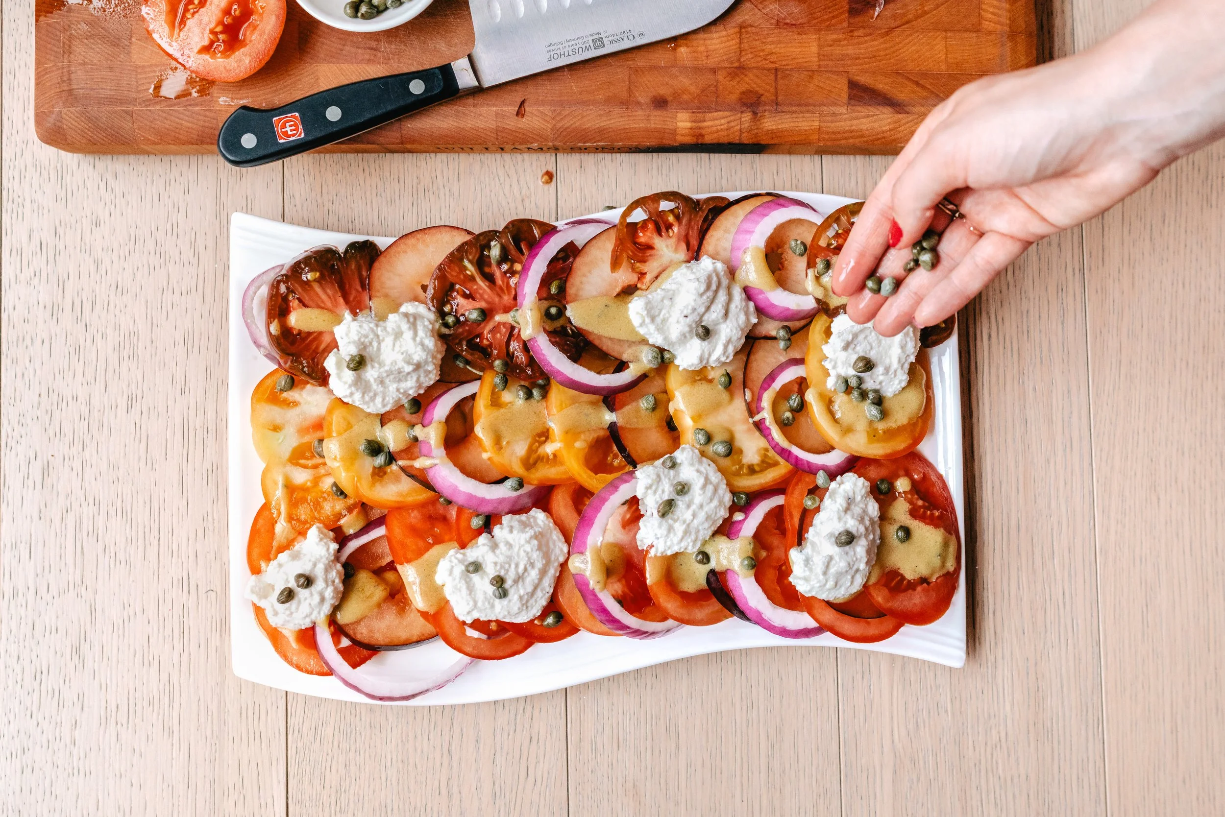 A food blogger and chef preparing a white rectangular plate of sliced heirloom tomatoes with dollops of white cheese and a hand sprinkling capers for their branding photoshoot.