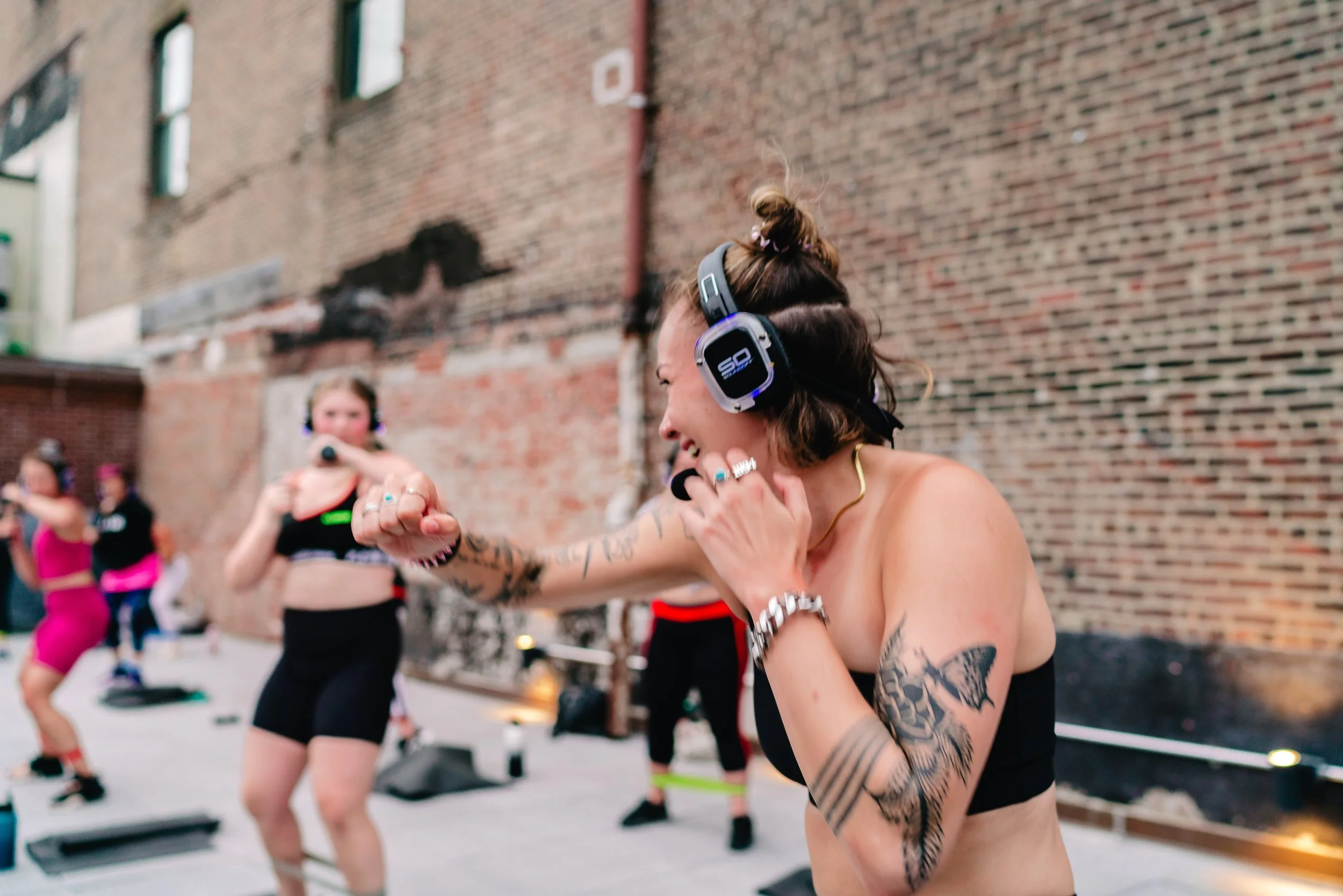 Women dancing and exercising outdoors against a brick wall, wearing headphones and fitness attire.