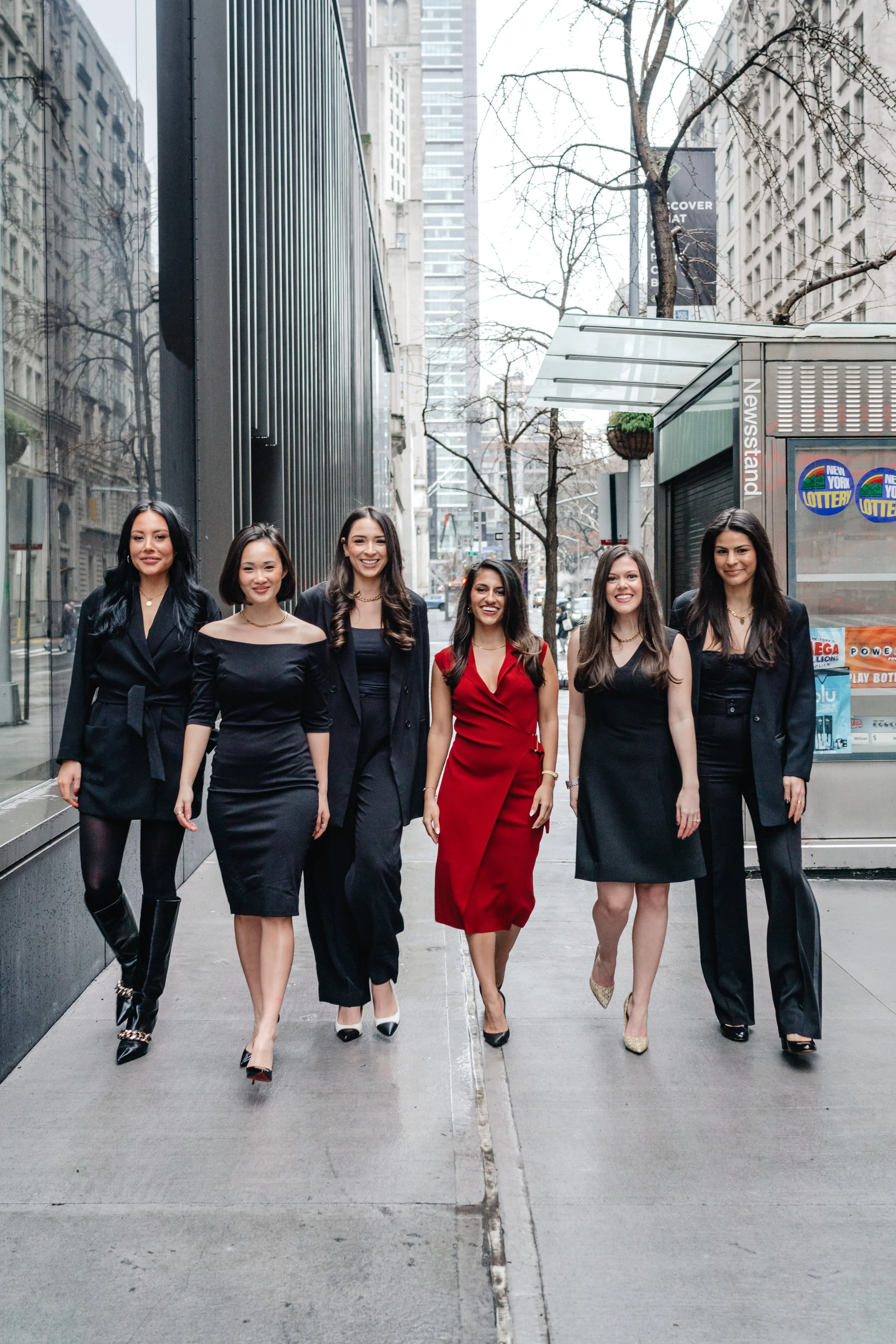 A group of six women lawyers walking together on a New York City sidewalk, dressed in fashion-forward black and red outfits, smiling and posing for the camera for their all female firm's brand photography photoshoot.