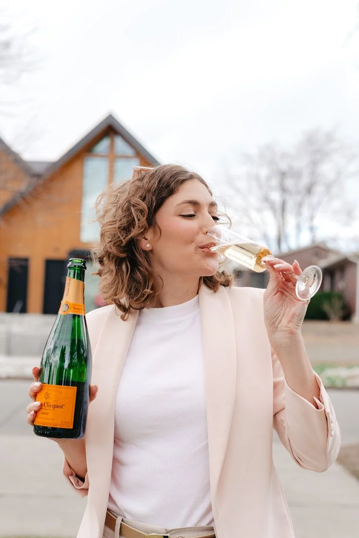 Woman with curly hair in a white shirt and beige blazer drinking champagne from a flute glass outdoors, holding a champagne bottle in her hand.