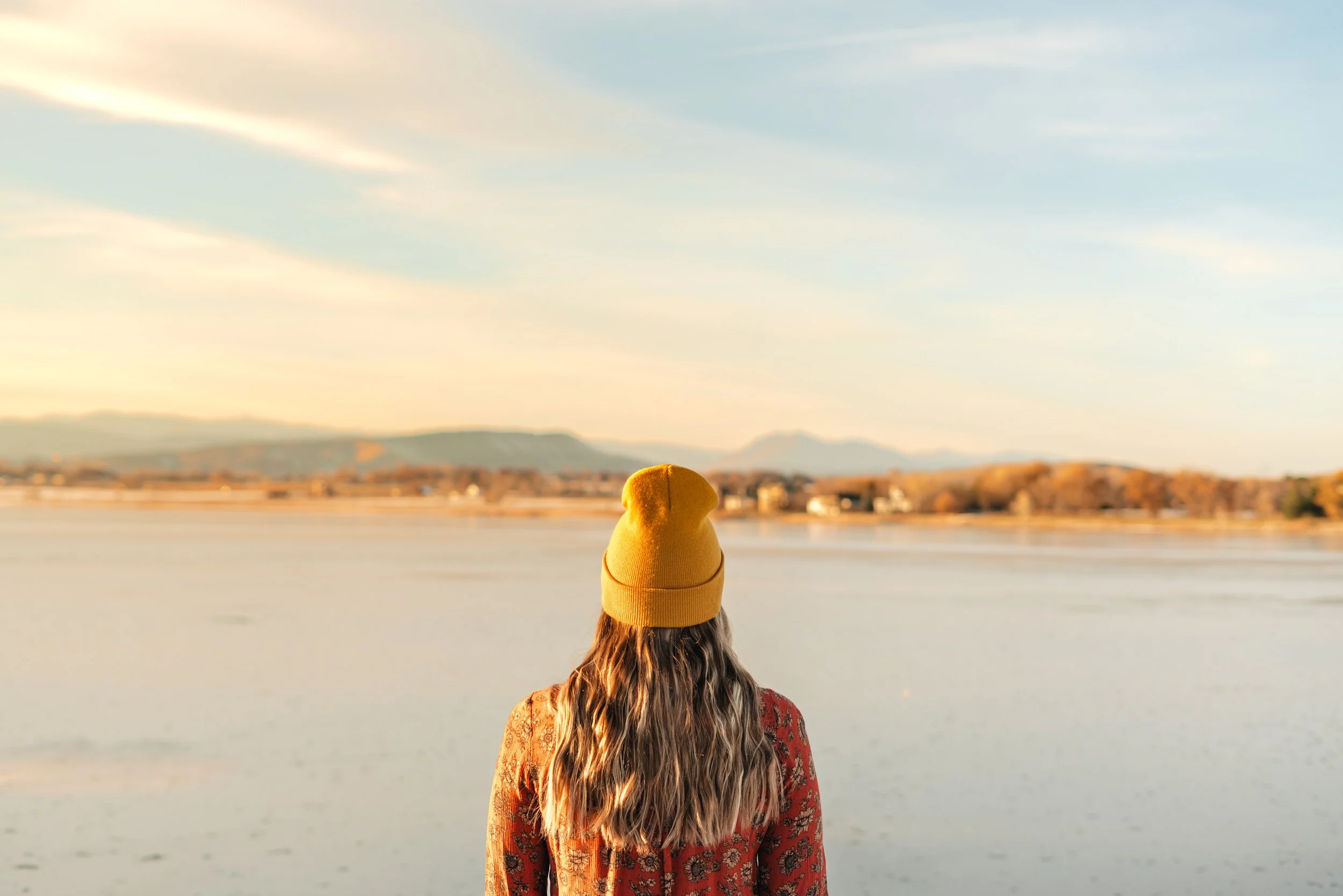 A woman wearing a yellow beanie and a patterned red sweater stands by a body of water at sunset, facing away from the camera.