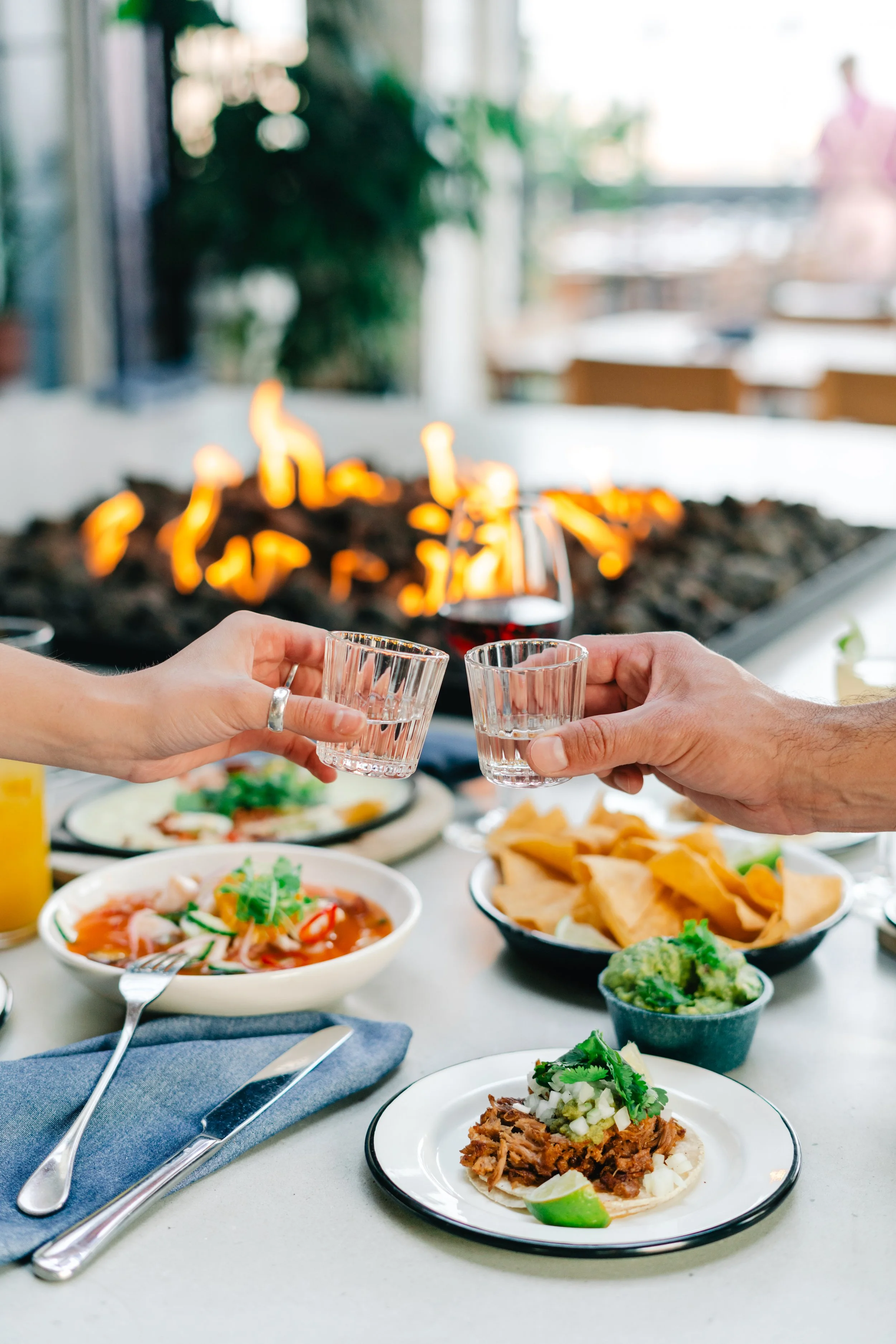 People clinking glasses of water at a table with Mexican food, including tacos, chips, guacamole, and salsa, with a flame feature in the background.