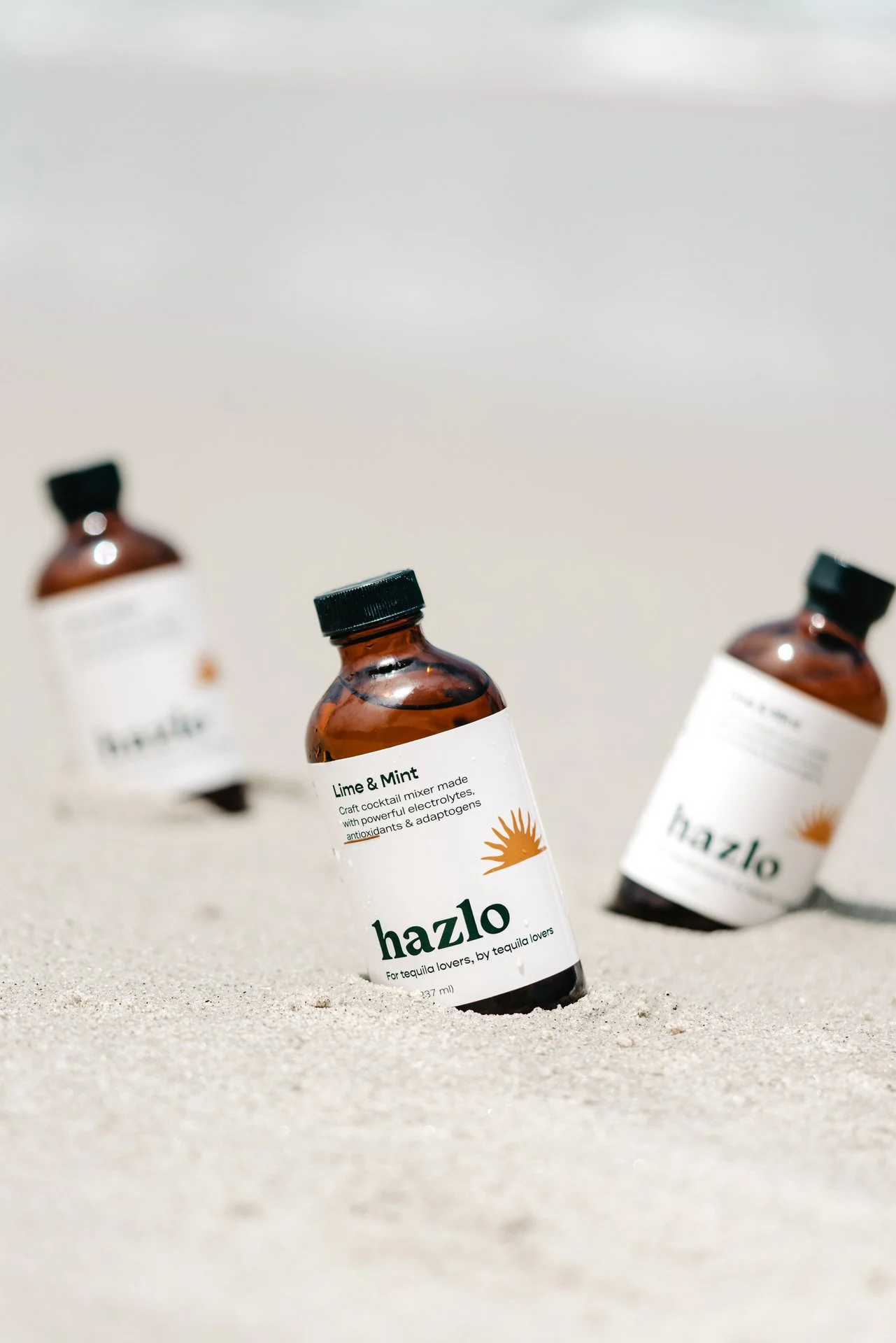 A product photo of three brown glass bottles labeled 'hazlo' with a sun logo, placed on sandy ground on a sandy Diego beach, with one bottle in focus and the other two blurred in the background.
