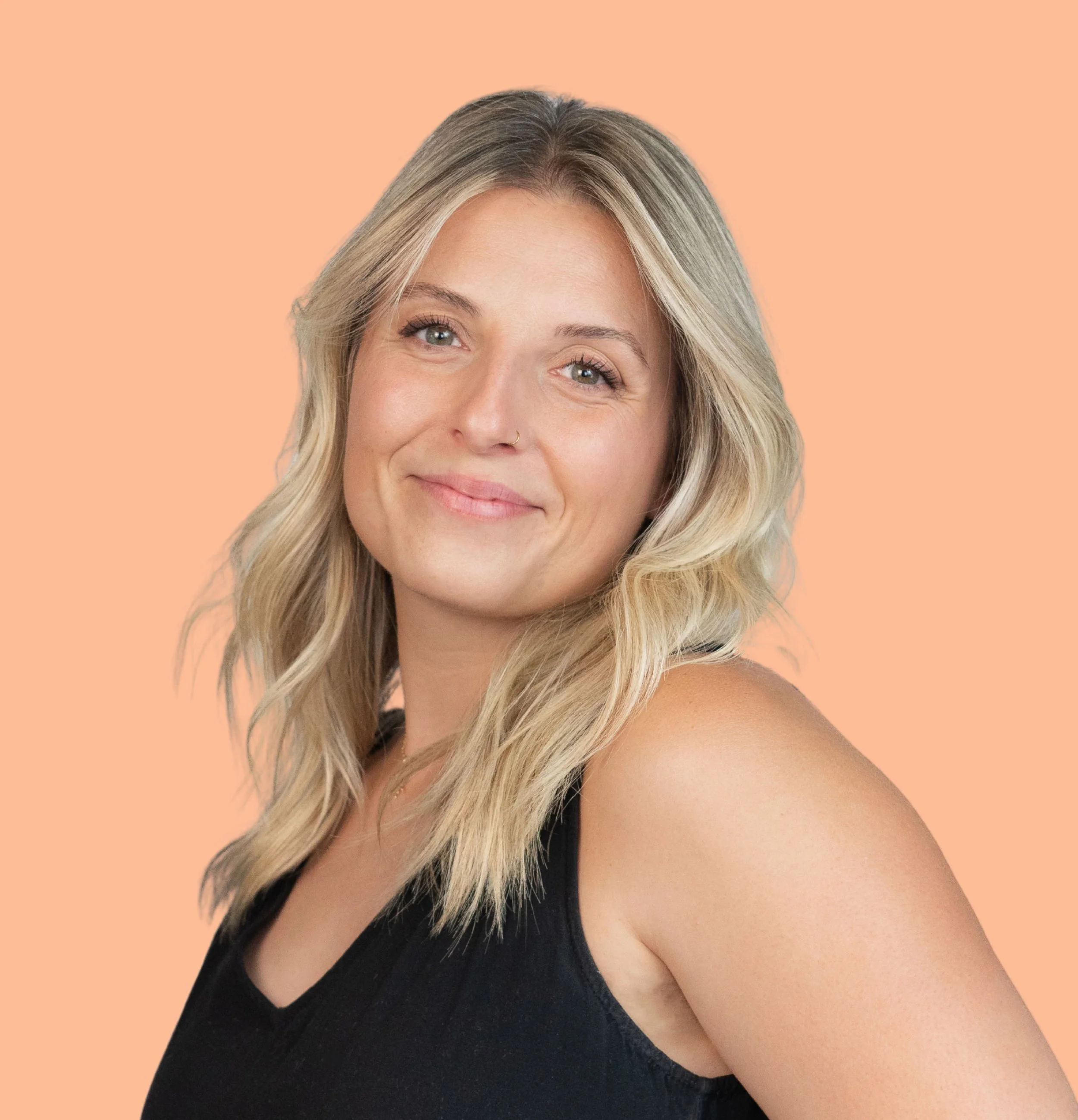 A young woman with blonde hair and a nose piercing smiling at the camera against a peach-colored background.