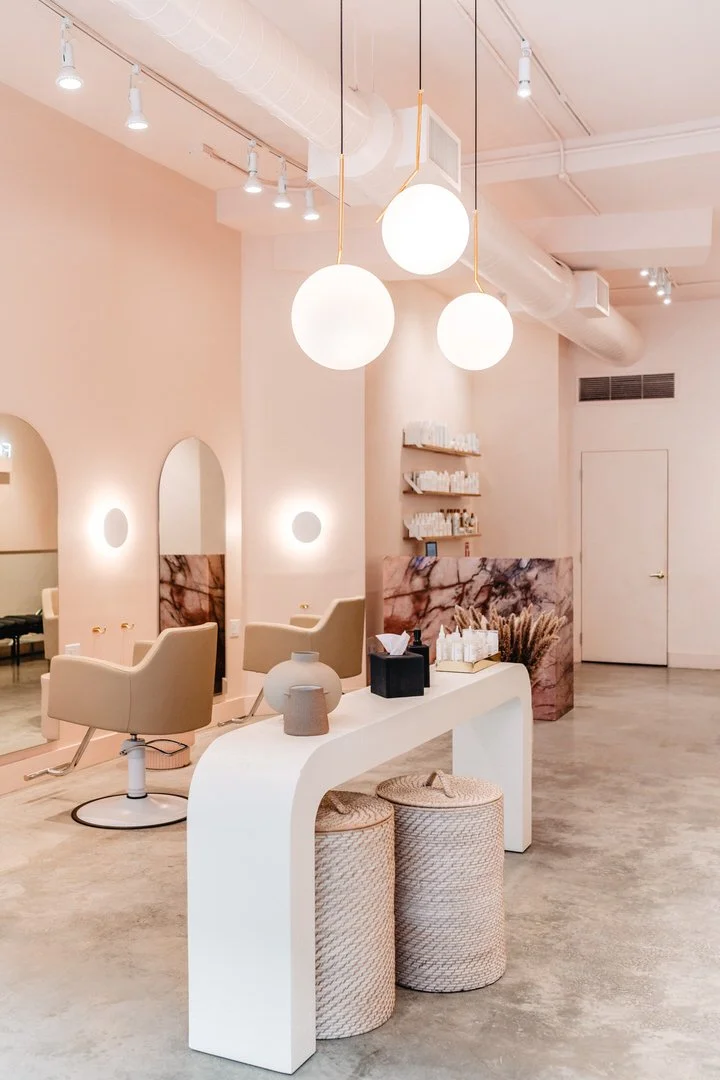 Interior of a modern salon with beige chairs, mirrors, and soft lighting, featuring a white table with decorative vases and textured stools in the foreground, and a reception area with shelves and a pink marble counter in the background.