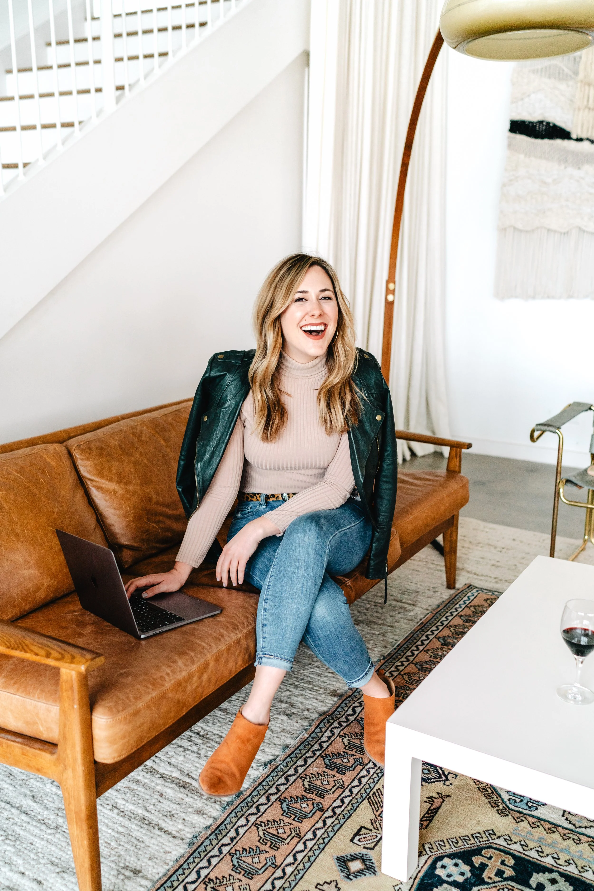 A woman sitting on a leather couch, smiling and laughing, with a laptop on her lap in a modern living room.