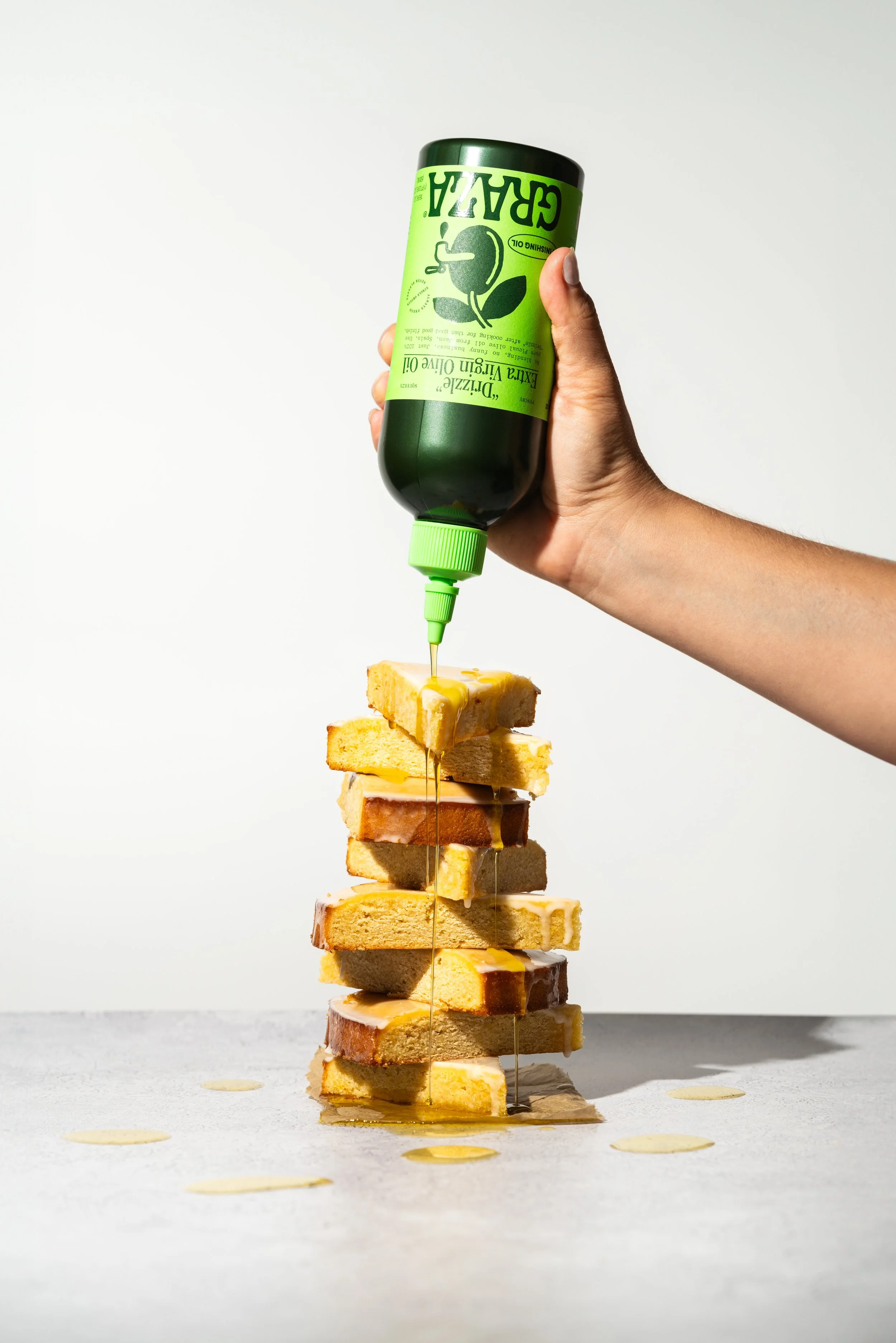 A hand holding a green bottle of olive oil poured over a stack of sliced bread on a white surface.