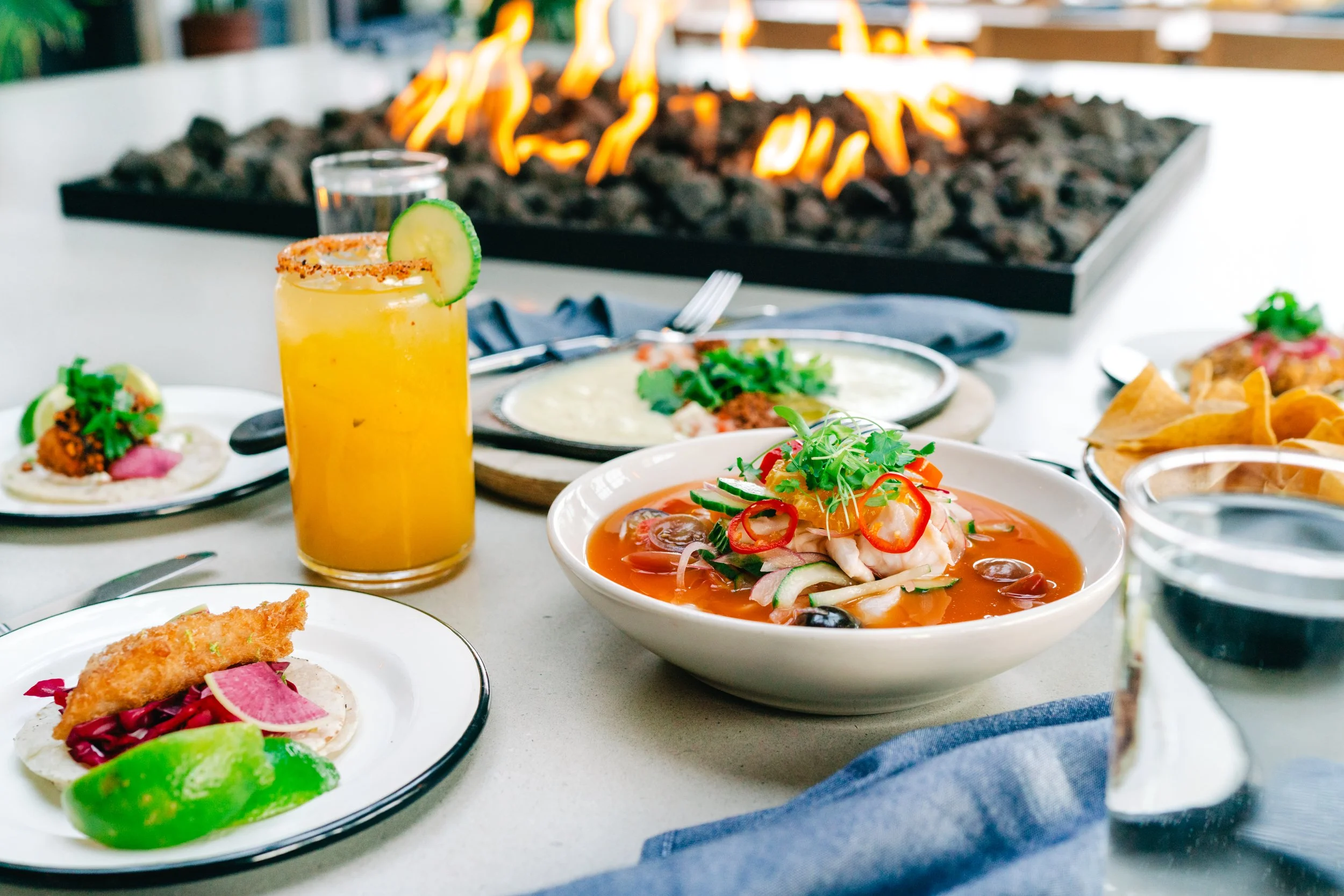 A table set with Mexican food and beverages, featuring a fire pit in the background. The dishes include a spicy margarita, a bowl of seafood soup, tacos with seafood and vegetables, a plate of fish tacos with lime and pickled vegetables, and a side of tortilla chips.