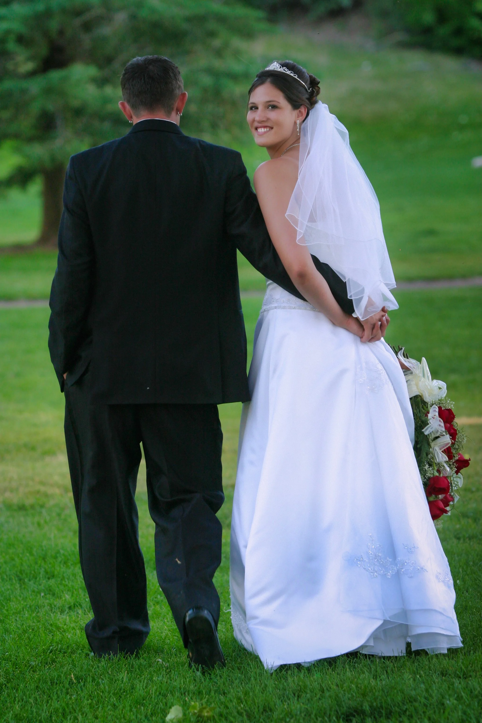 Bride and groom holding hands in a park setting, with the bride smiling at the camera, wearing a white wedding dress and veil, and the groom in a black suit