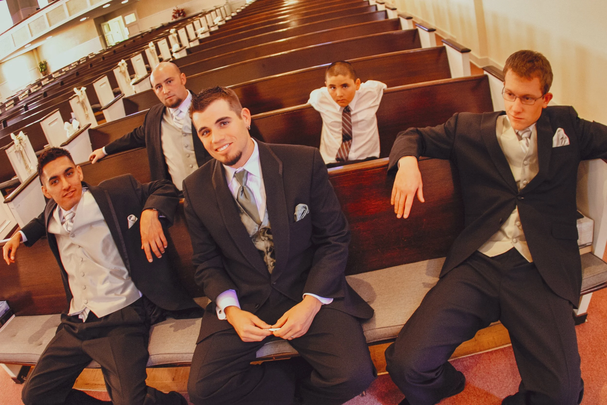 Group of five young men in formal suits sitting in pews inside a church or chapel.