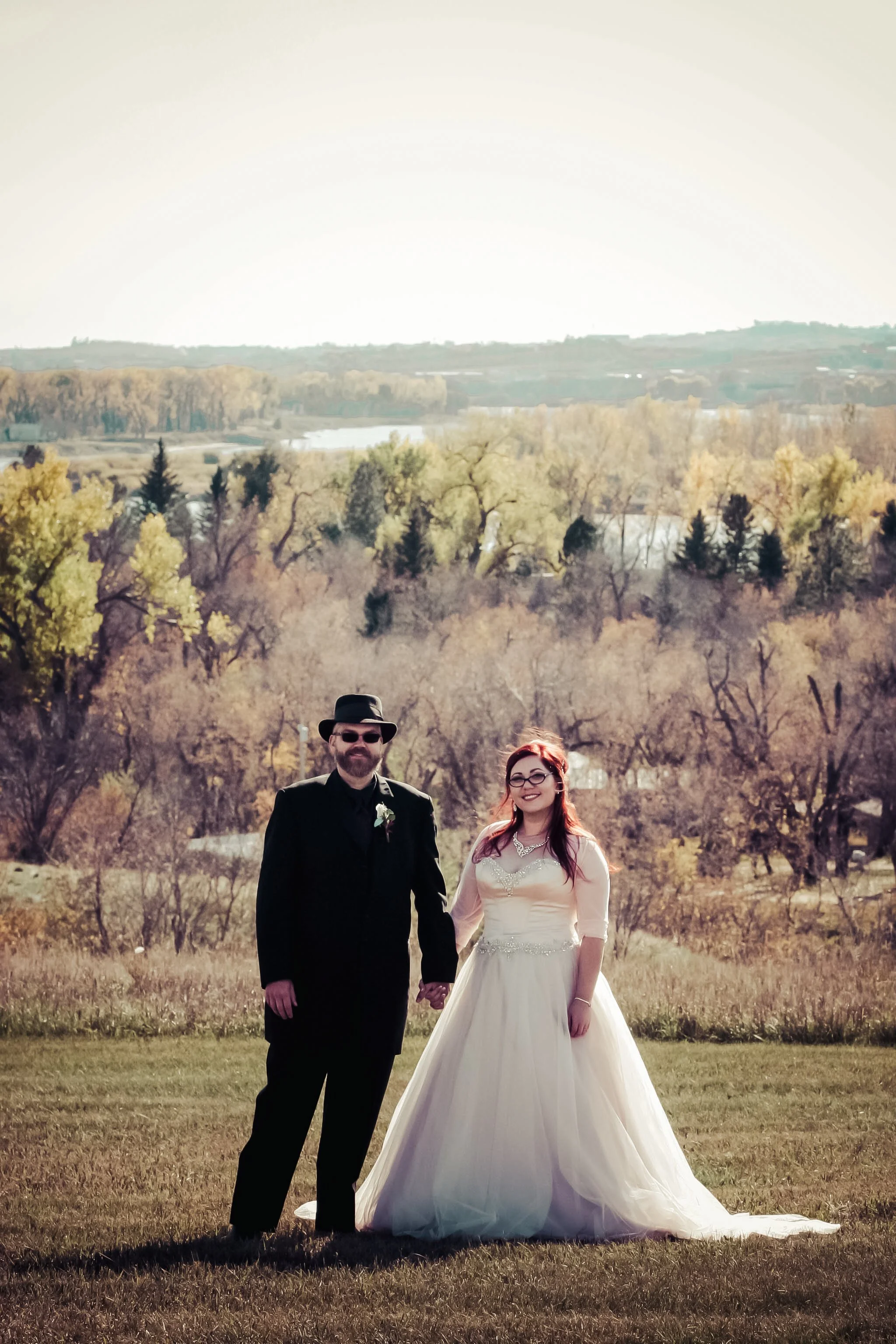 A bride and groom holding hands outdoors on a grassy area with trees and hills in the background. The groom is wearing a black suit, tie, and a black hat and sunglasses. The bride is wearing a white wedding dress with lace and beading, and glasses. T