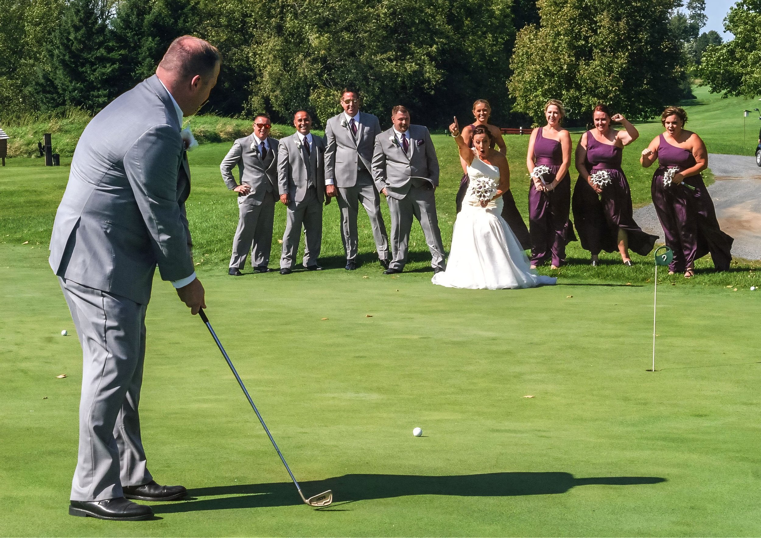 A man in a gray suit playing golf on a green with a group of women and men in formal attire watching and smiling, wedding party in the background.
