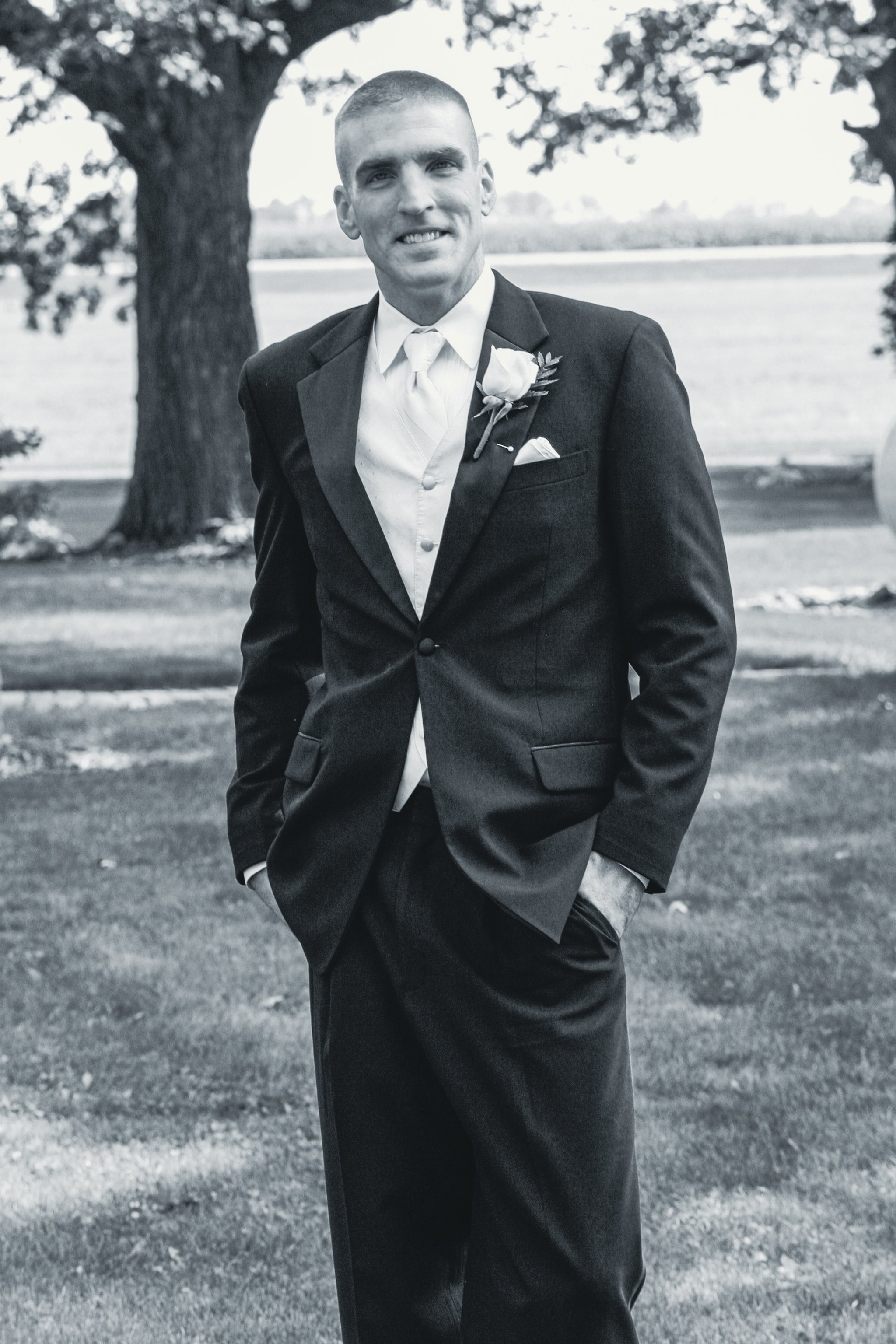 A man wearing a tuxedo standing outdoors with trees and a body of water in the background, smiling with hands in his pockets.