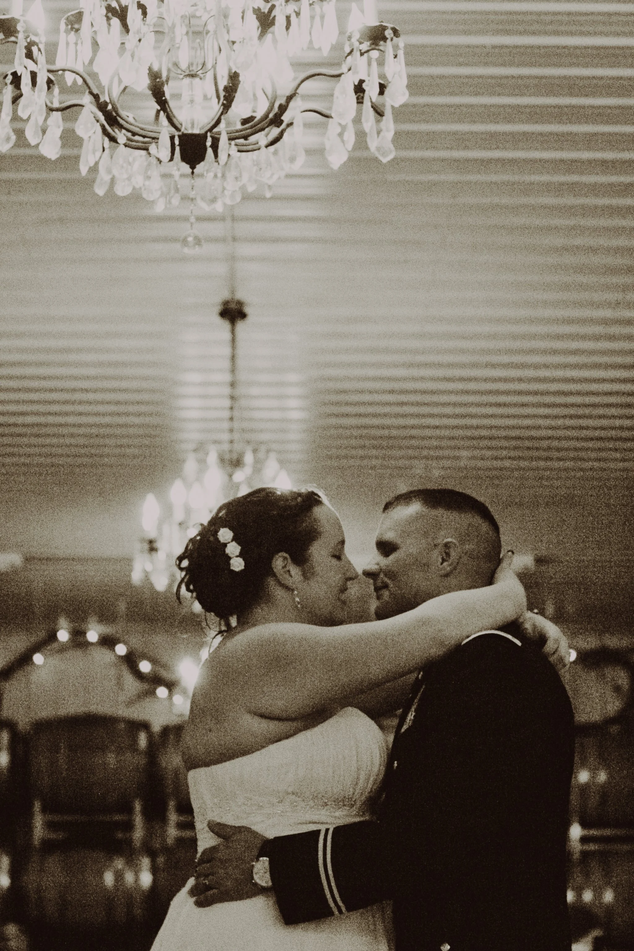 A black and white photo of a bride and groom sharing a dance at their wedding reception, with a chandelier hanging above them.