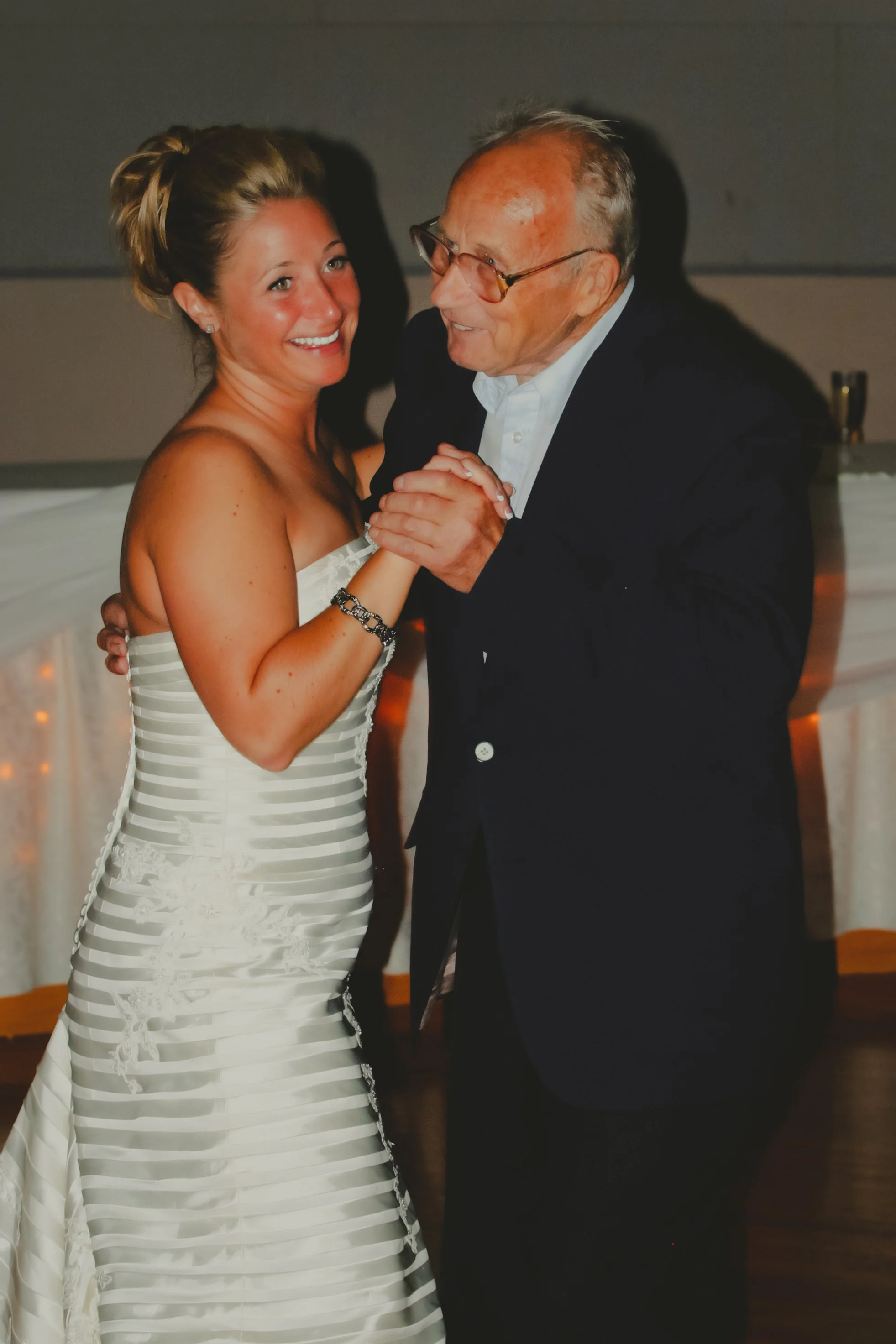 A young woman in a white wedding dress dancing with an older man in a black suit at a wedding reception.