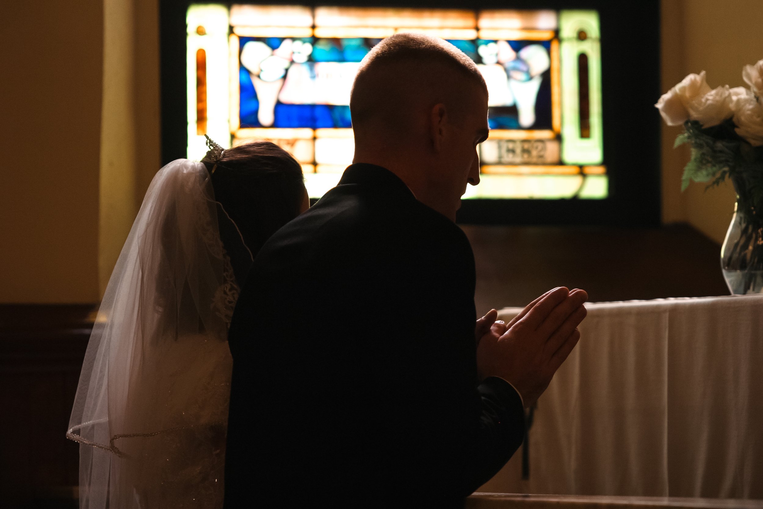 A man and a woman kneeling in prayer at a wedding ceremony, with a stained glass window behind them and a vase of white flowers nearby.