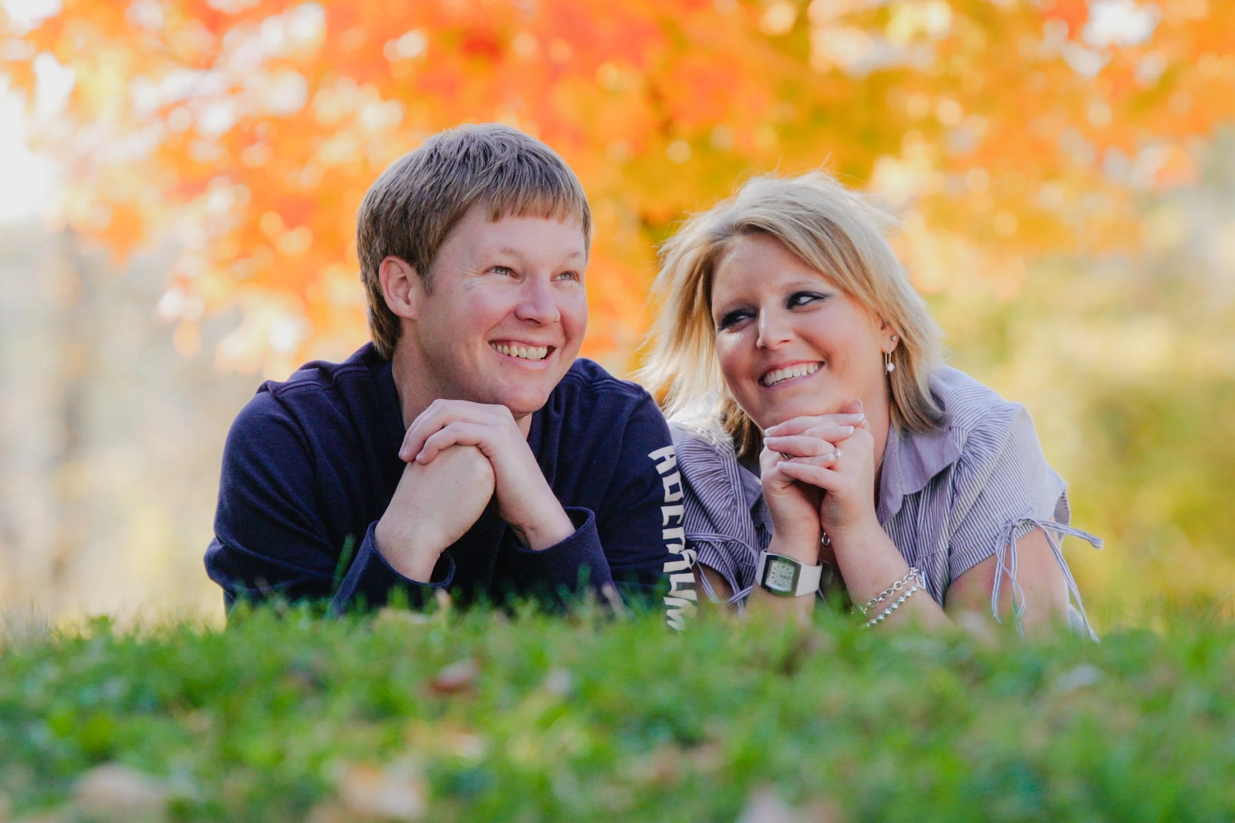 A young man and woman are lying on the grass outdoors in autumn, smiling and looking at each other. The background features colorful fall foliage with orange and yellow leaves.