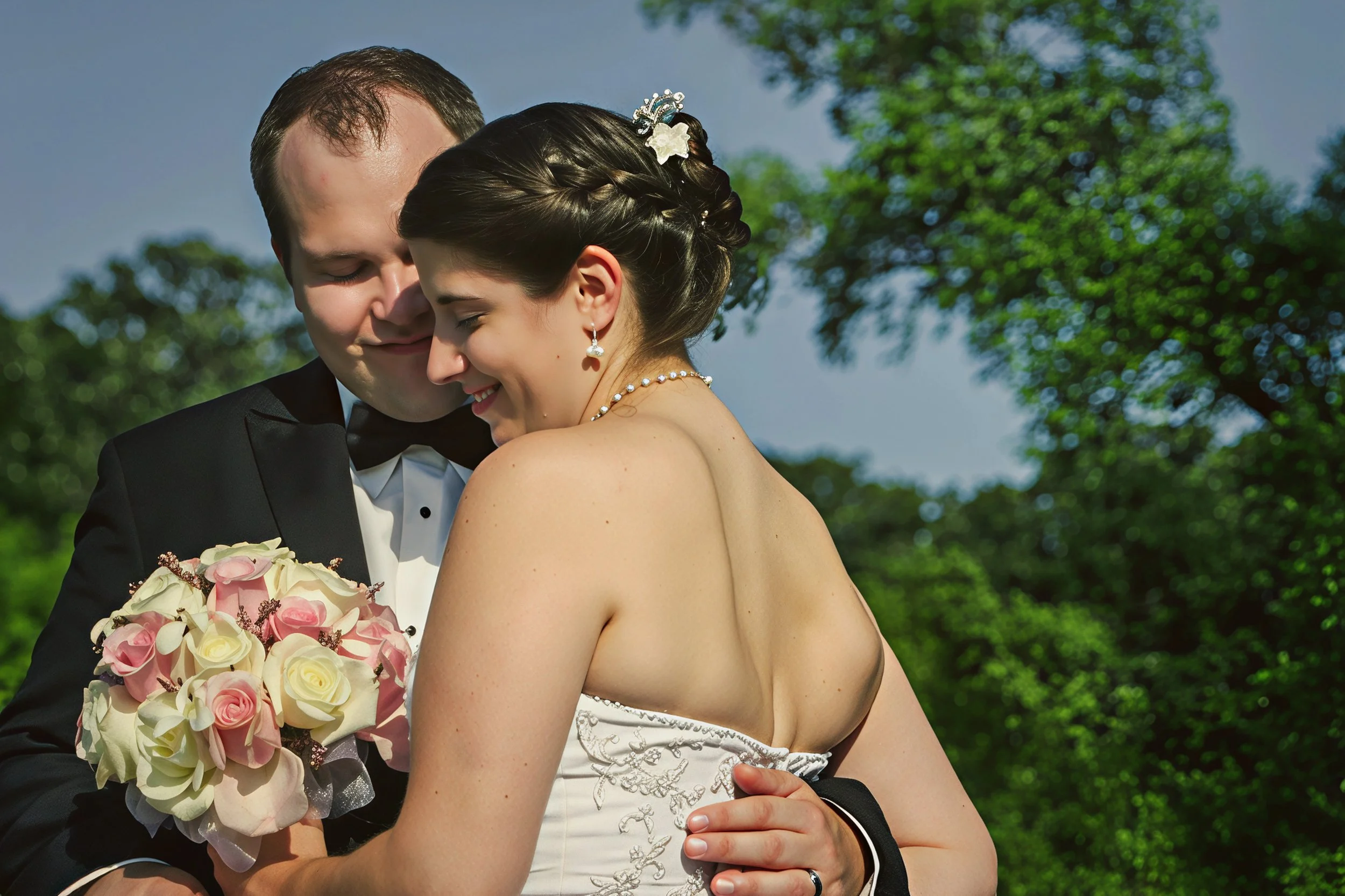 A groom and bride embrace and smile with closed eyes, outdoors under a blue sky and green trees. The bride holds a bouquet of pink and white roses, and wears a white strapless wedding gown with embroidered details, pearl earrings, and a pearl necklac