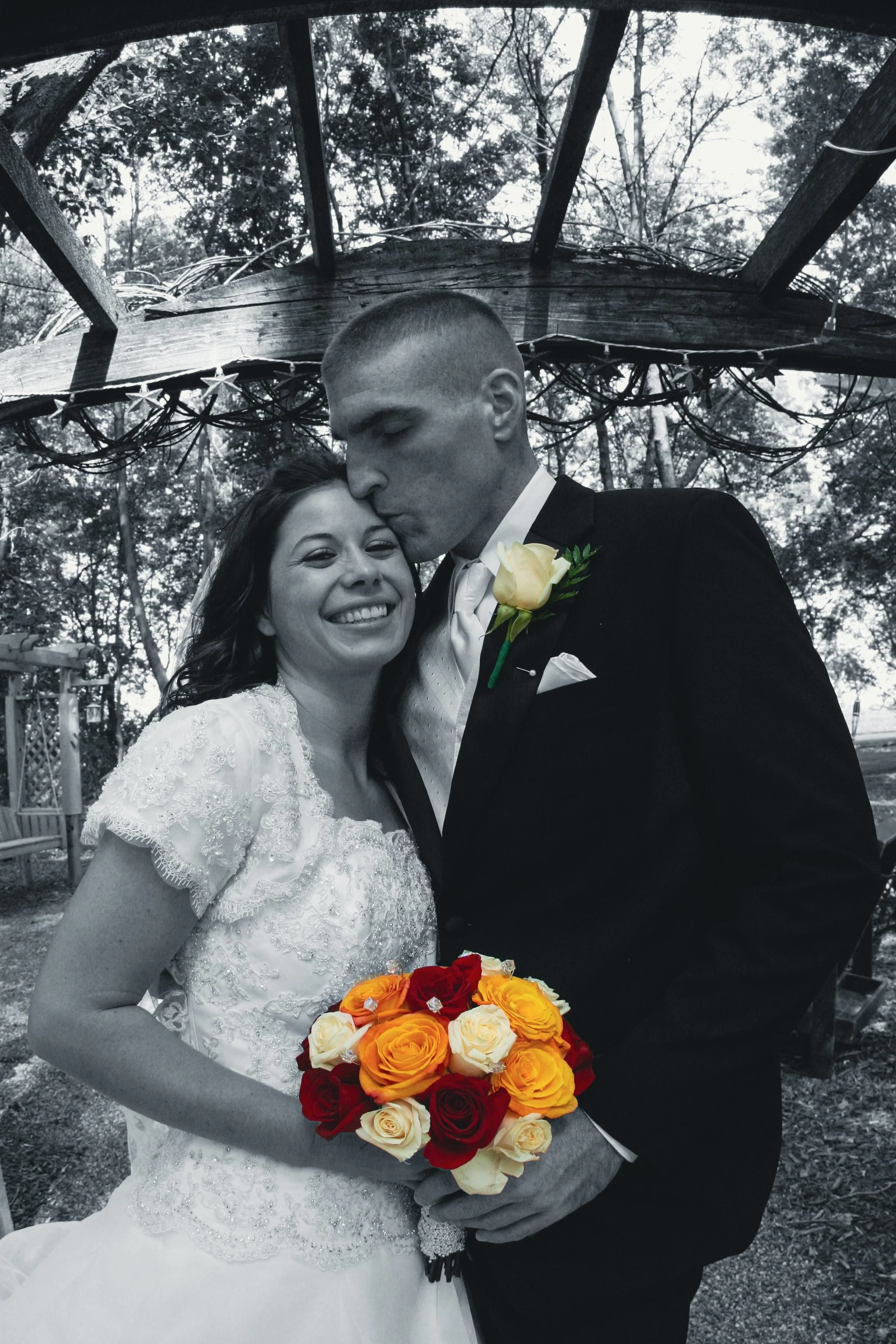 A black and white photo of a bride and groom. The groom, in a dark suit with a yellow boutonniere, kisses the bride on the forehead. The bride, in a lace wedding dress, smiles holding a colorful bouquet of orange, red, and cream roses. They are outdo