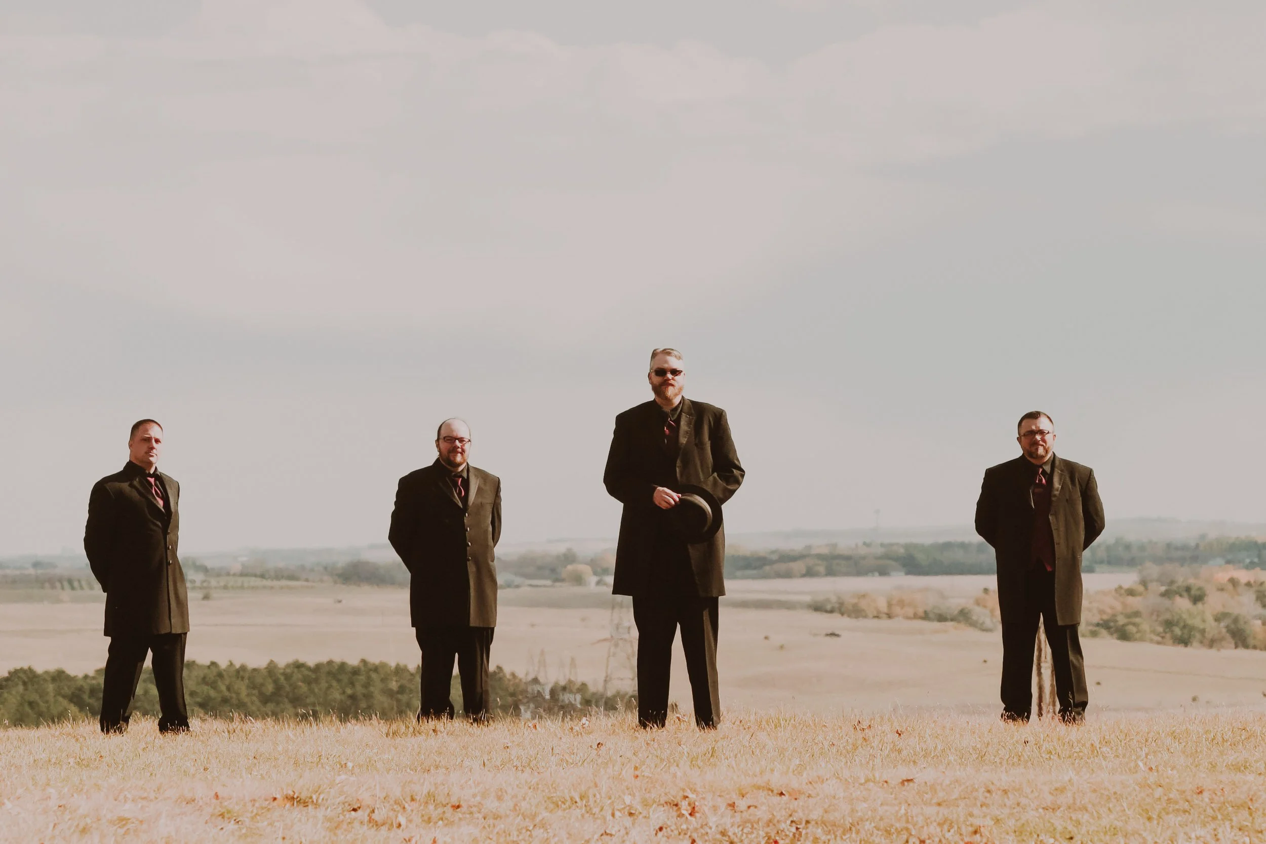 Four men dressed in dark suits and ties standing on a grassy plain outdoors with a vast landscape and sky in the background. One man in the middle holds a hat.
