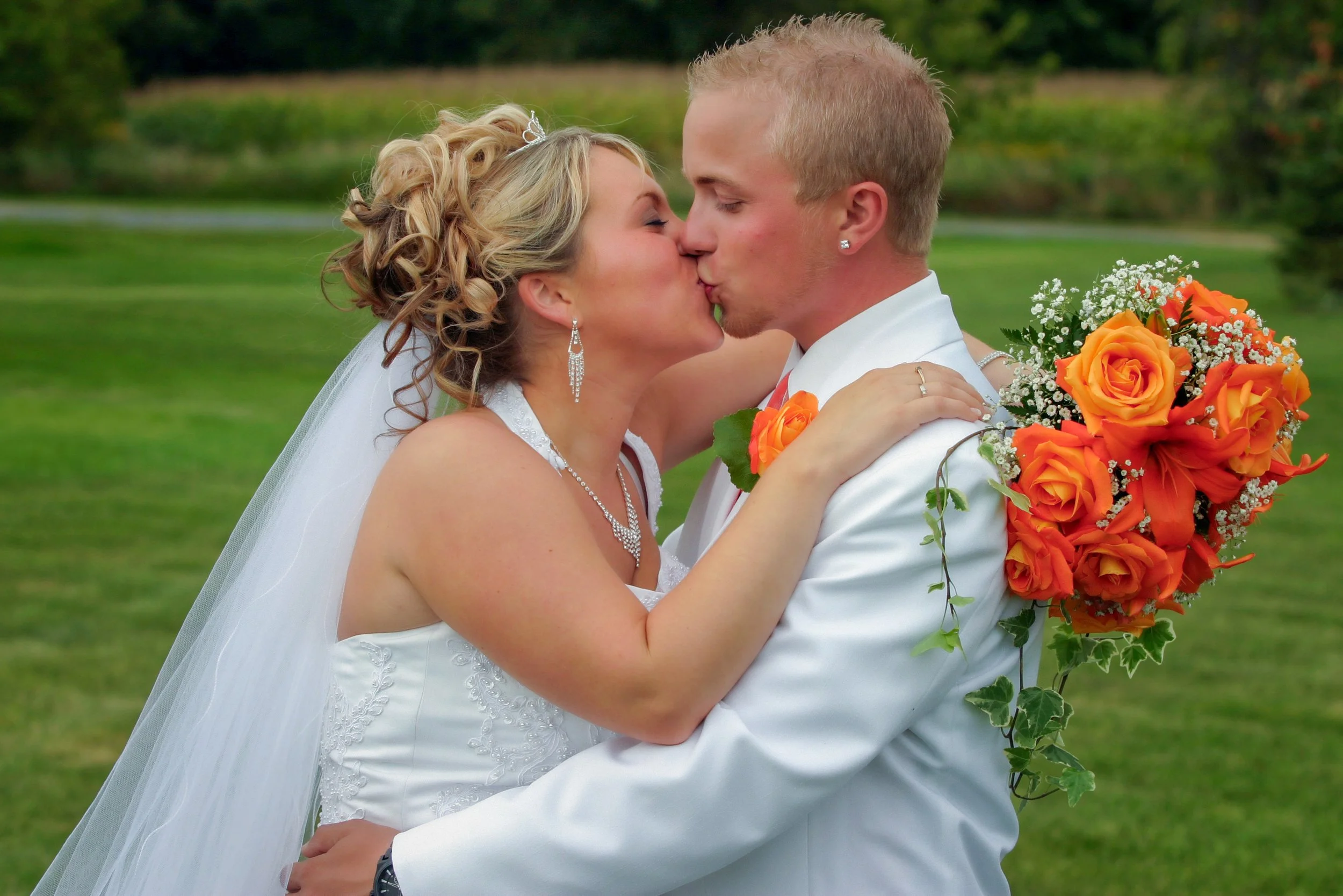 A bride and groom kiss outdoors on their wedding day, the bride holding a bouquet of orange roses.