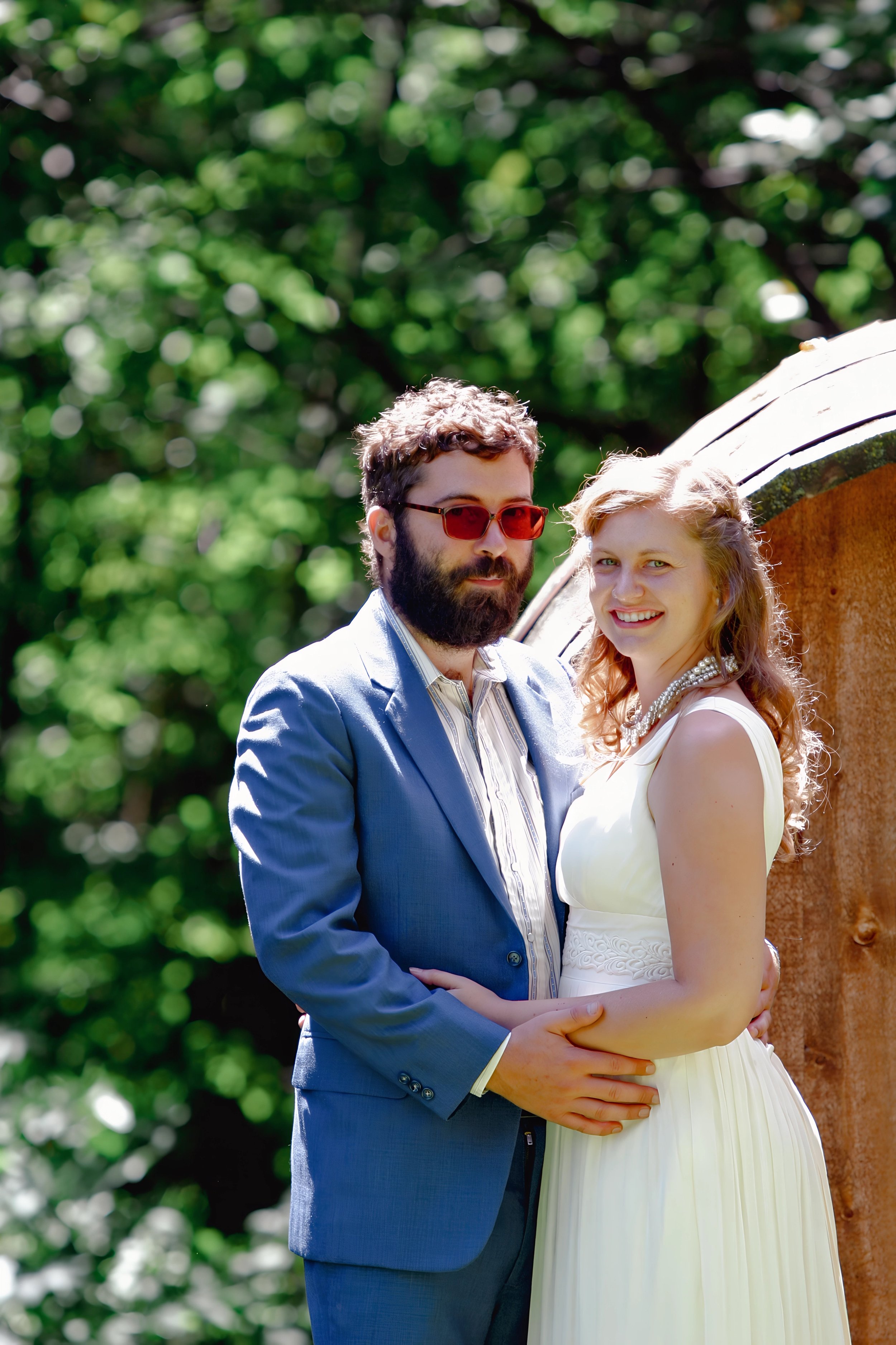 A man and woman standing outdoors, embracing each other; the man has a beard, sunglasses, and is wearing a blue suit; the woman has curly hair, and is wearing a white dress and pearl necklace; trees in the background.