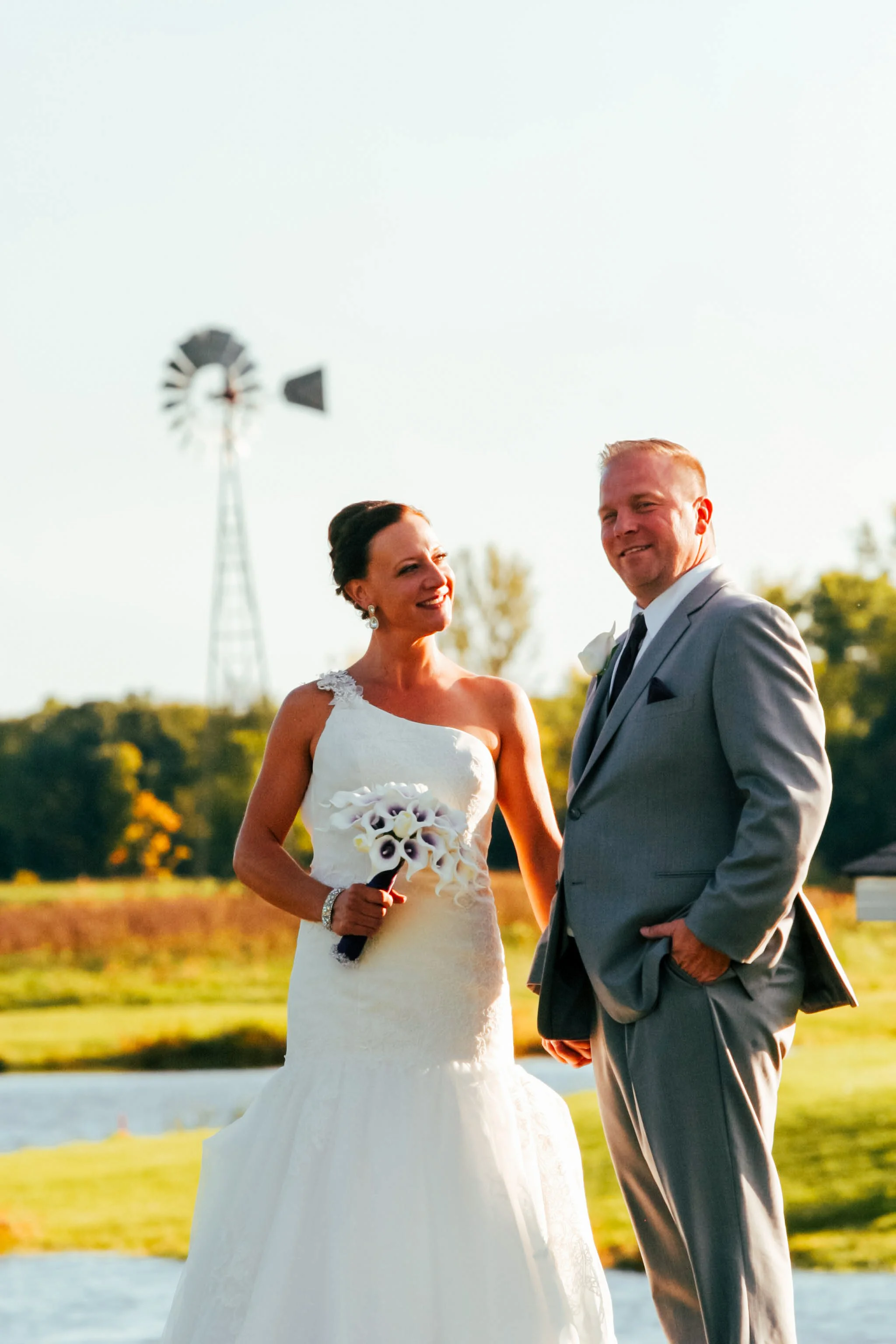 A bride and groom standing outdoors during their wedding, smiling at each other, with a windmill and trees in the background.