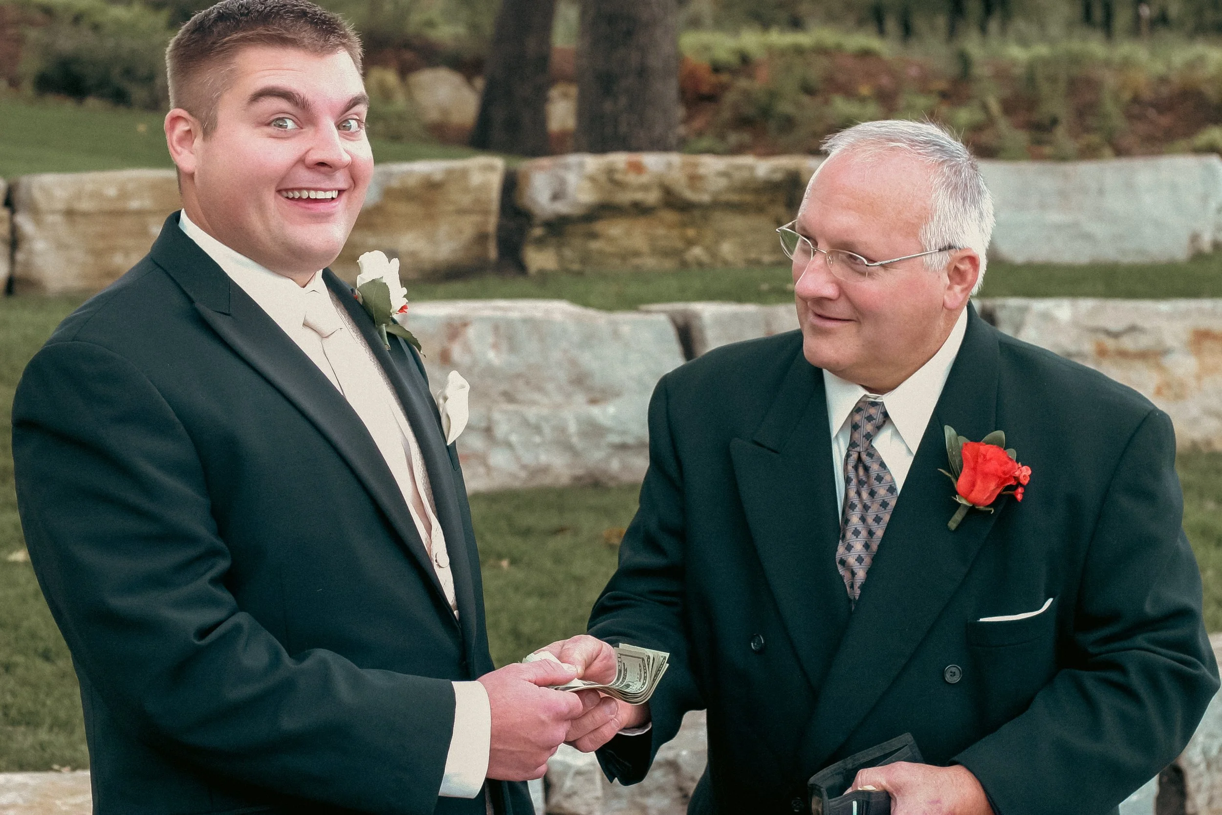 A young man in a tuxedo and an older man in a suit exchanging money during a wedding ceremony outdoors, with trees and rocks in the background.