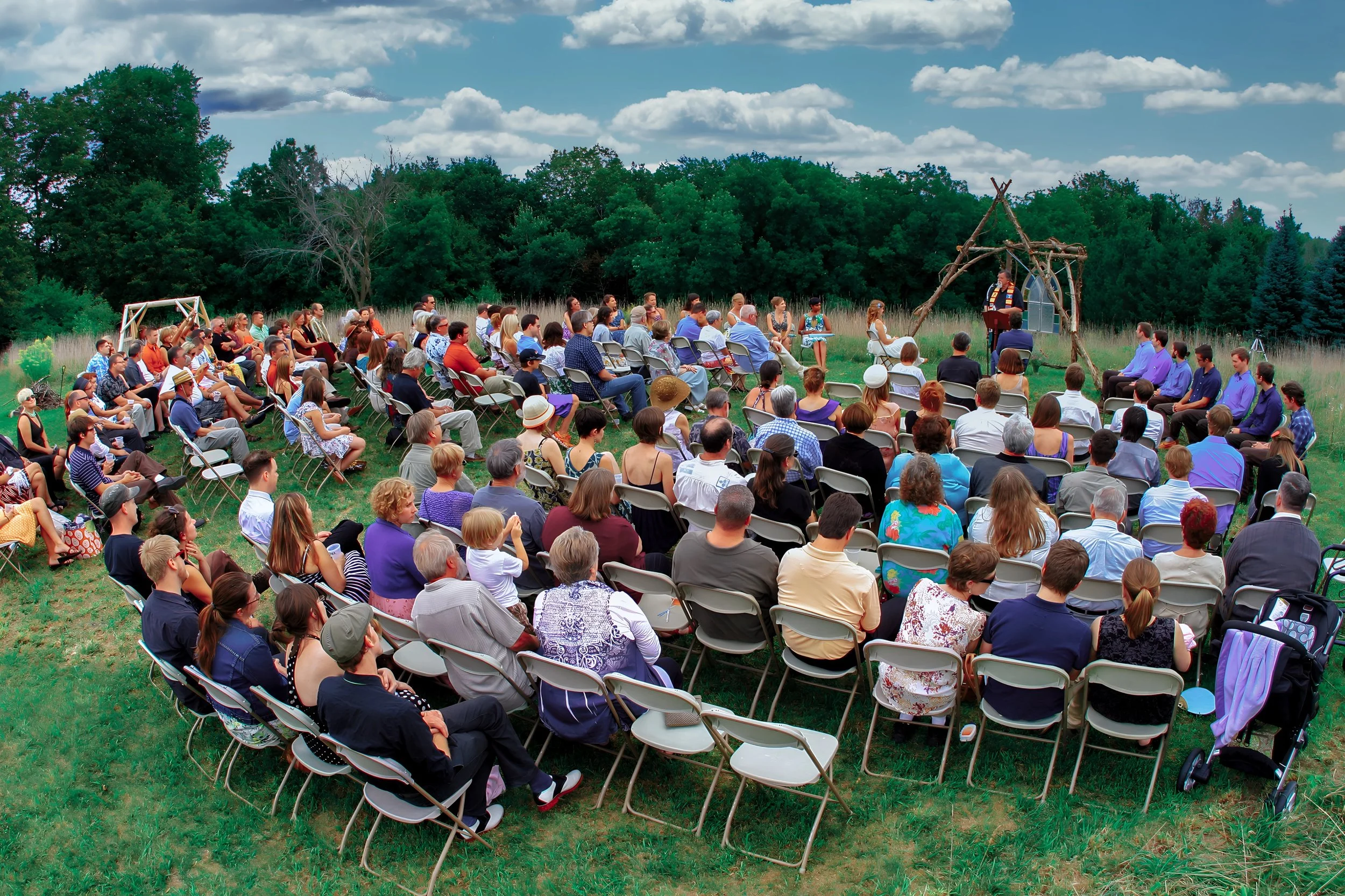 An outdoor gathering with many people seated on folding chairs listening to a speaker at a rustic stage in a grassy field, surrounded by trees and a partly cloudy sky.
