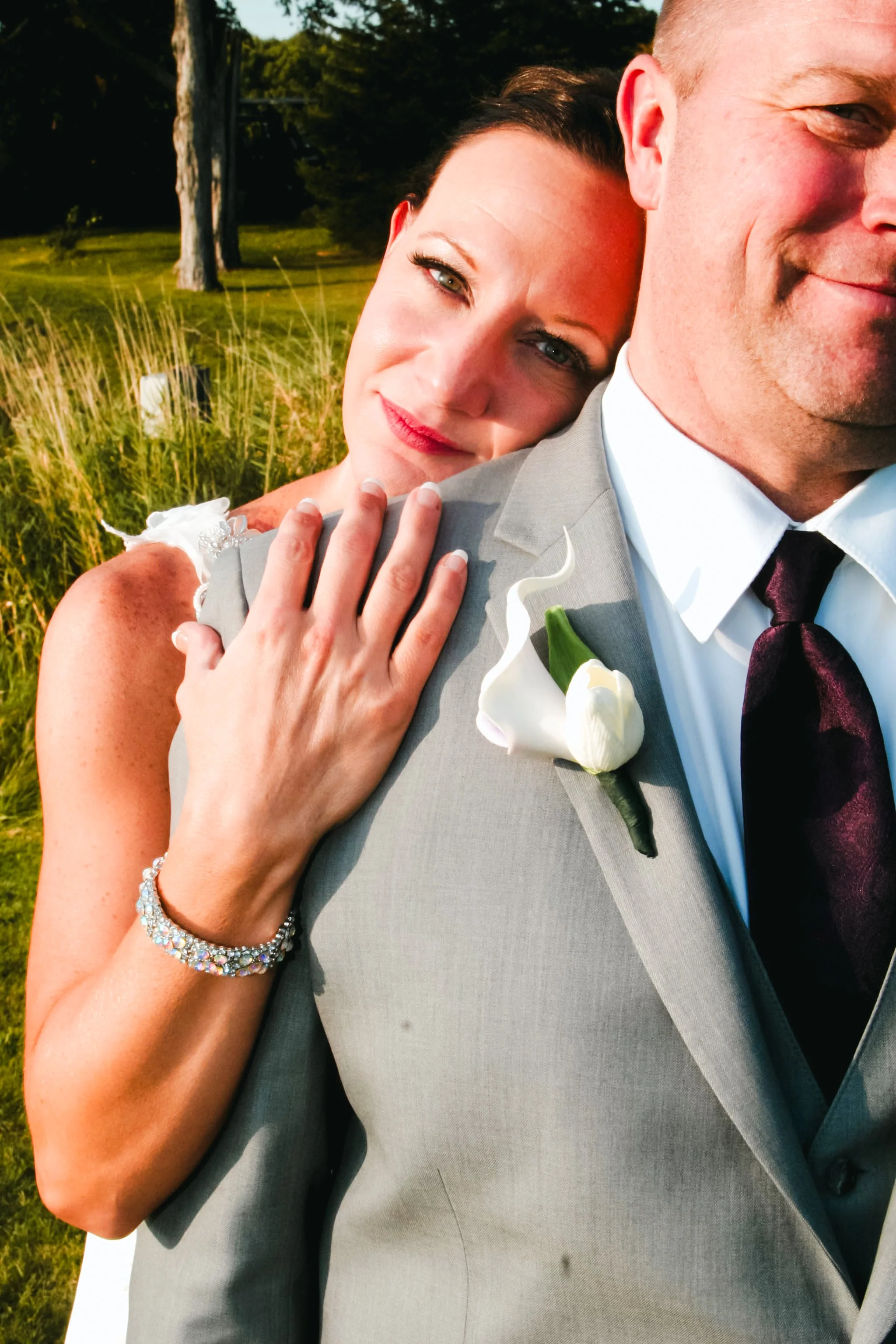 A woman with makeup and a bracelet, with her arm on a man's shoulder, hugging him from behind, outdoors near a grassy field, during a sunny day.