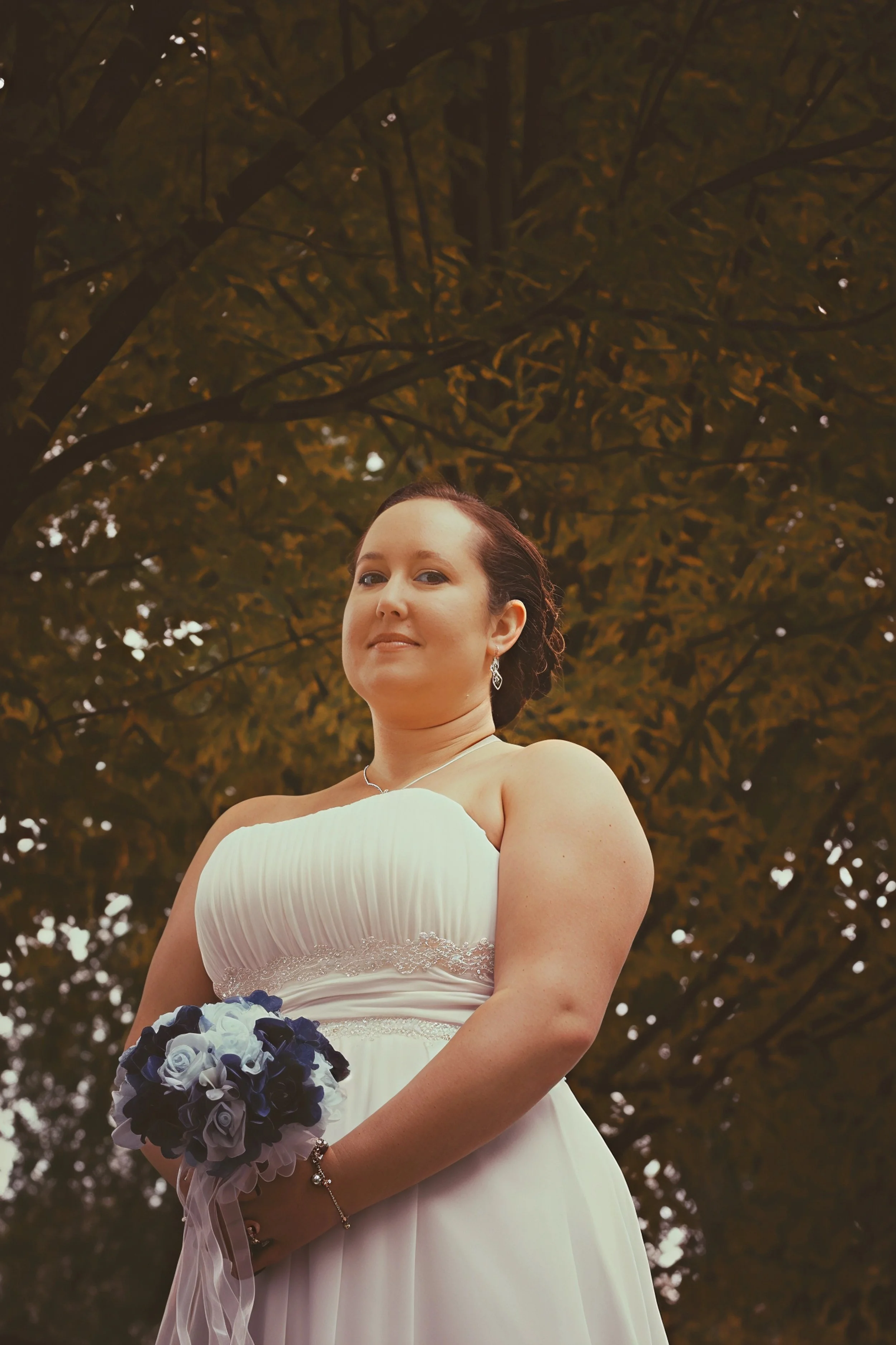 A bride in a white strapless wedding dress holding a bouquet of blue and white flowers, standing outdoors with a tree in the background.