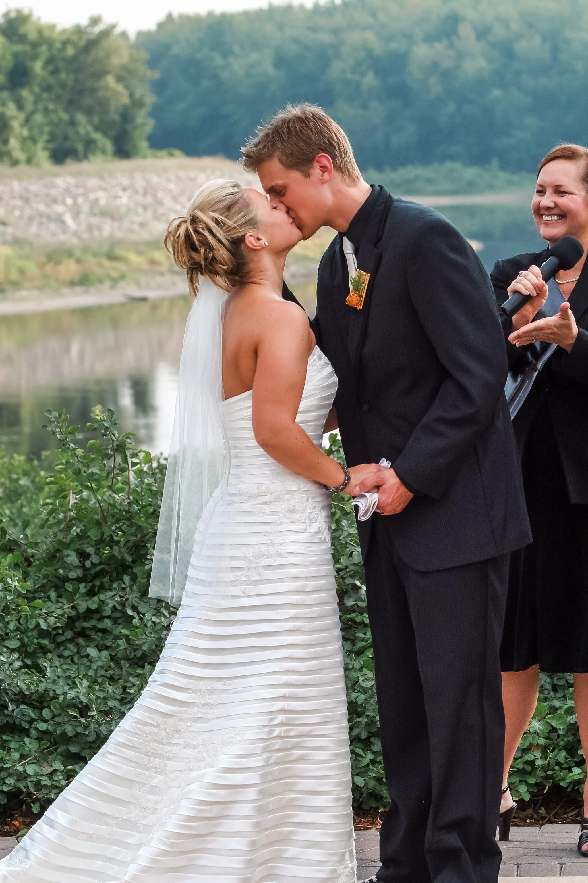 A bride and groom kiss during their outdoor wedding ceremony by a river, with a woman holding a microphone smiling in the background.