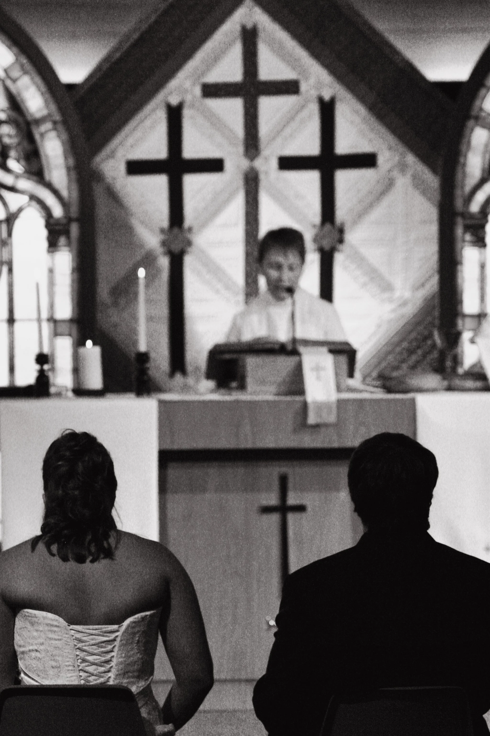 A bride and groom sit facing an altar in a church, watching a screen that displays a young boy speaking, with three crosses mounted on the wall behind him.