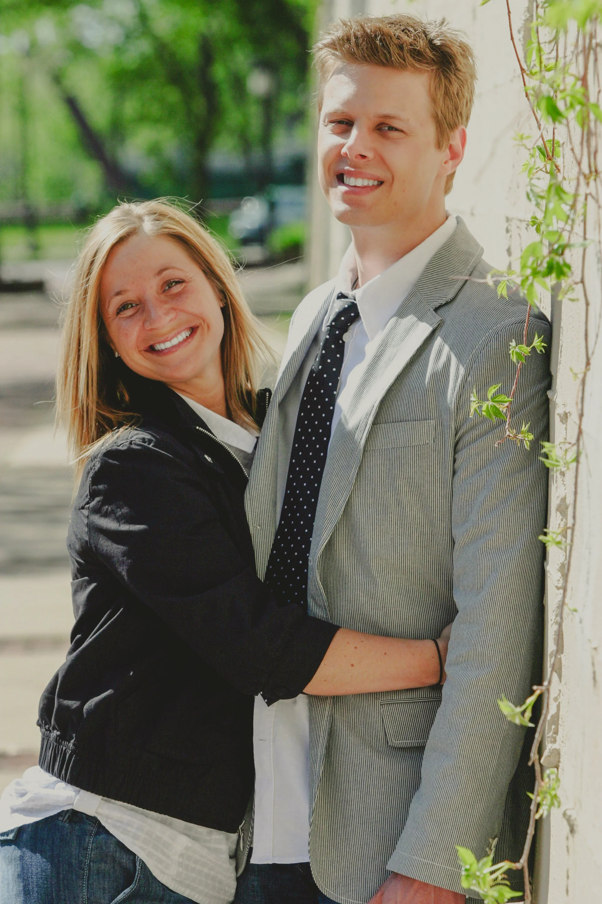 A smiling young woman and man embracing outdoors, standing against a wall with greenery in the background.